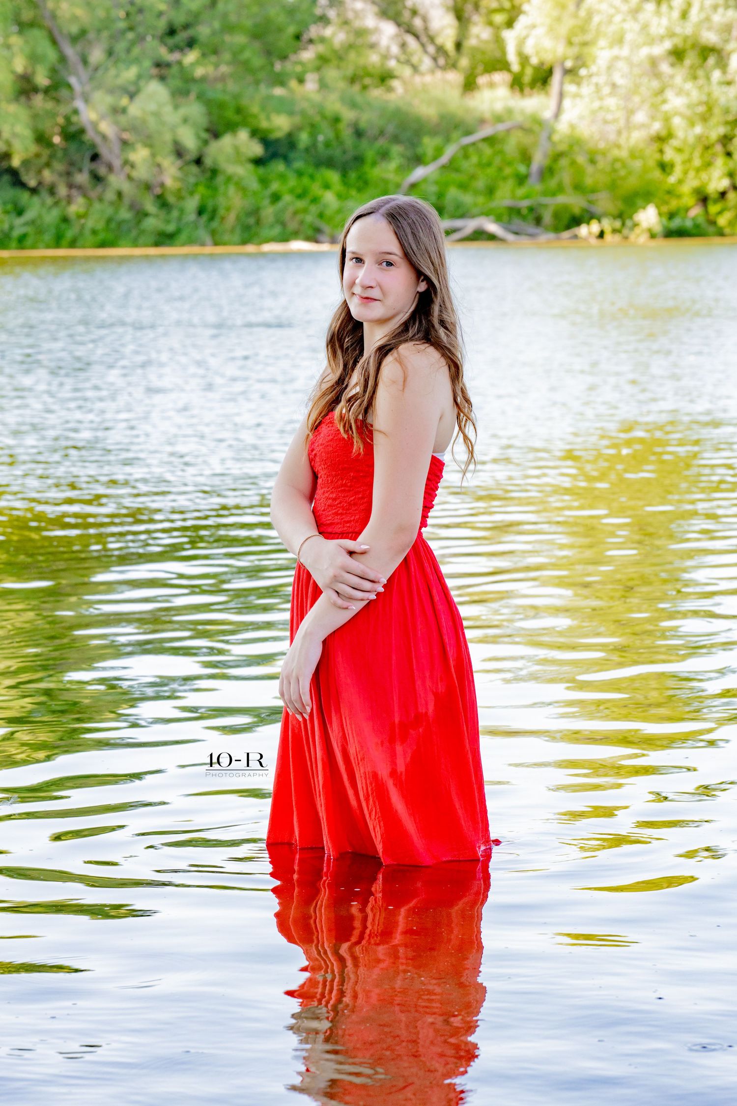 A young woman in a red gown stands in a calm lake, surrounded by lush greenery, posing for a portrait.