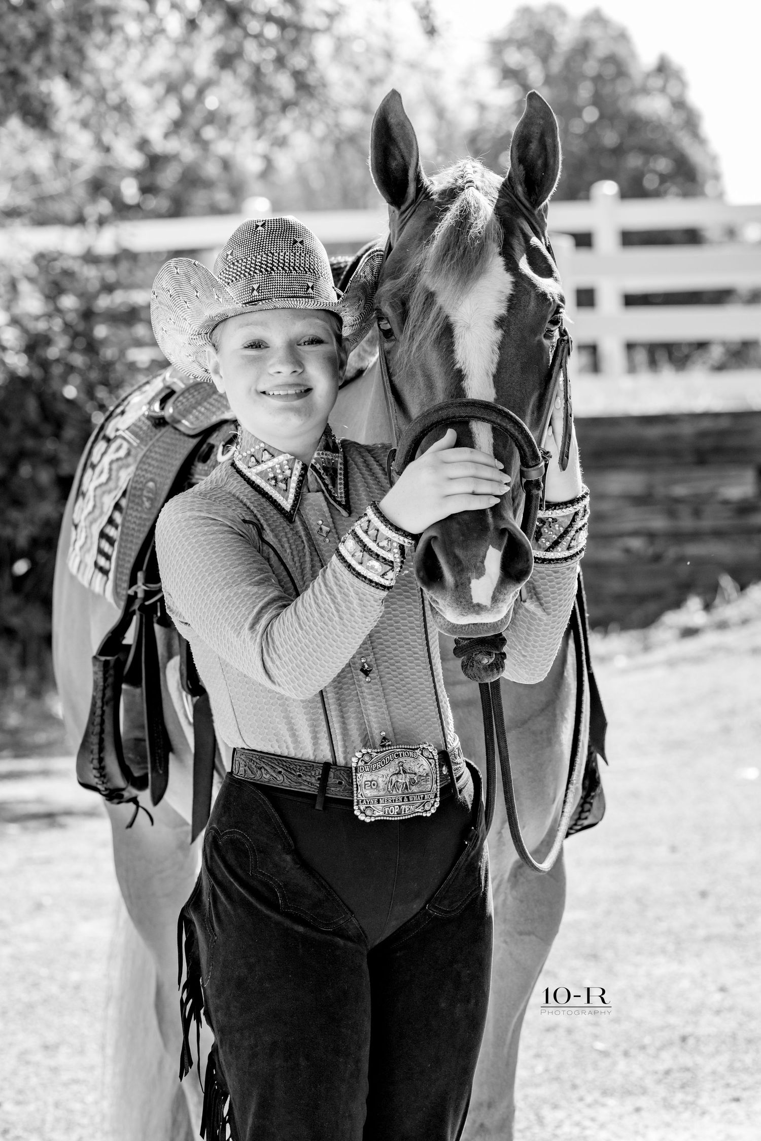 Black and white photo of a smiling young cowgirl in a decorative hat hugging her horse near a white fence.