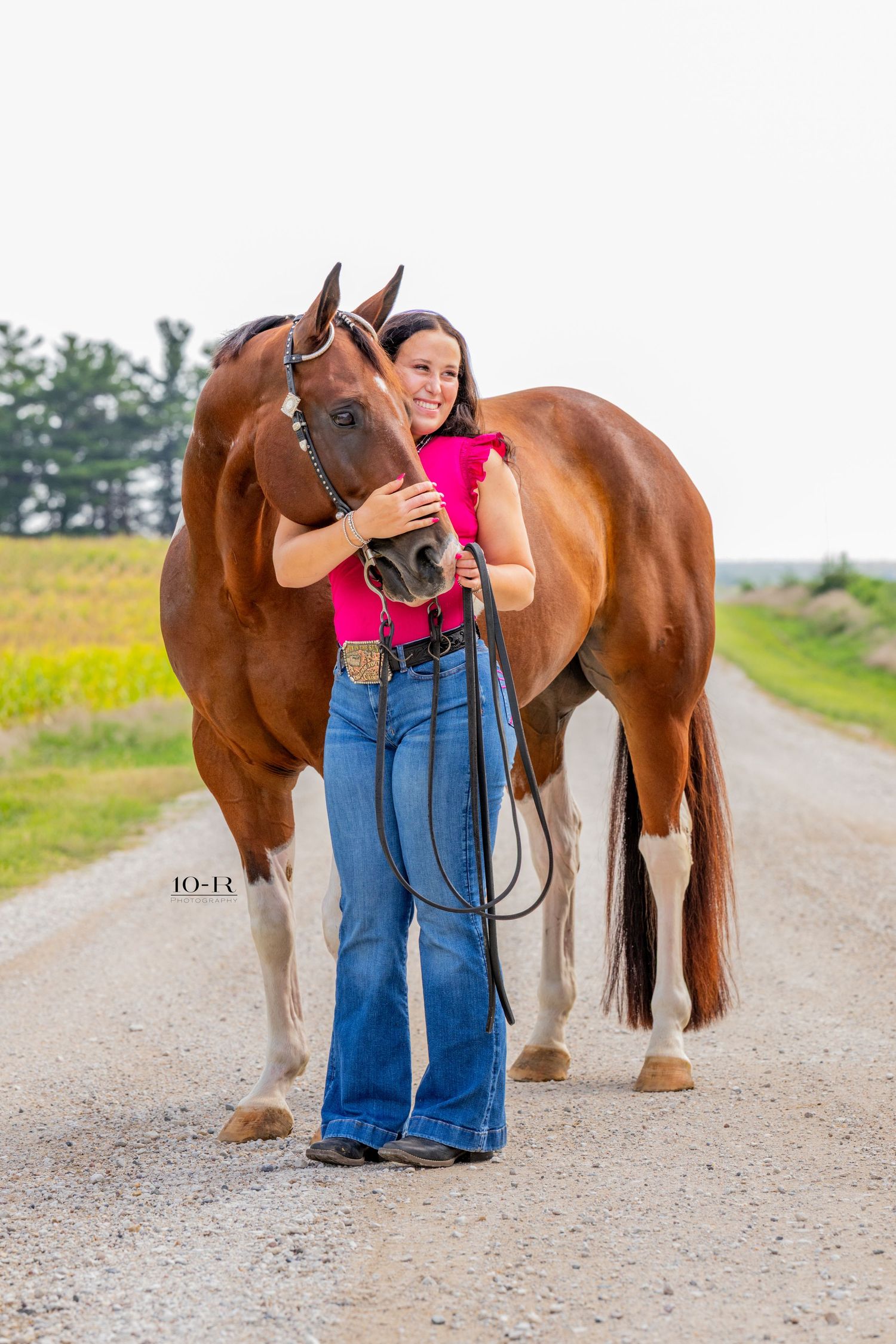 A smiling woman in a pink top and jeans hugs a chestnut horse on a rural gravel road surrounded by green fields.