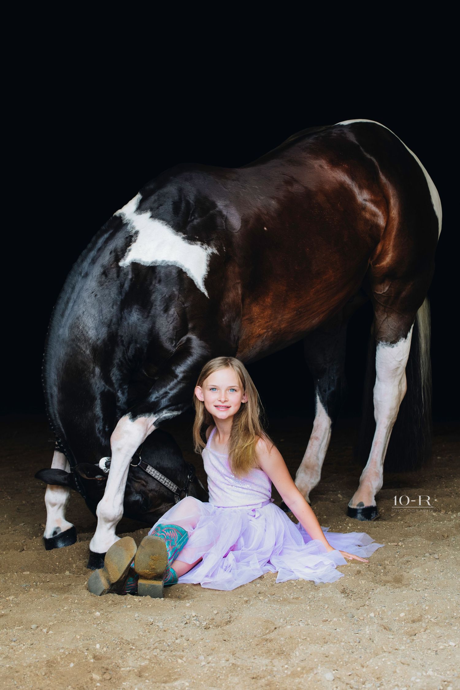 A child in a purple dress sits on sandy ground next to a large black and white horse.