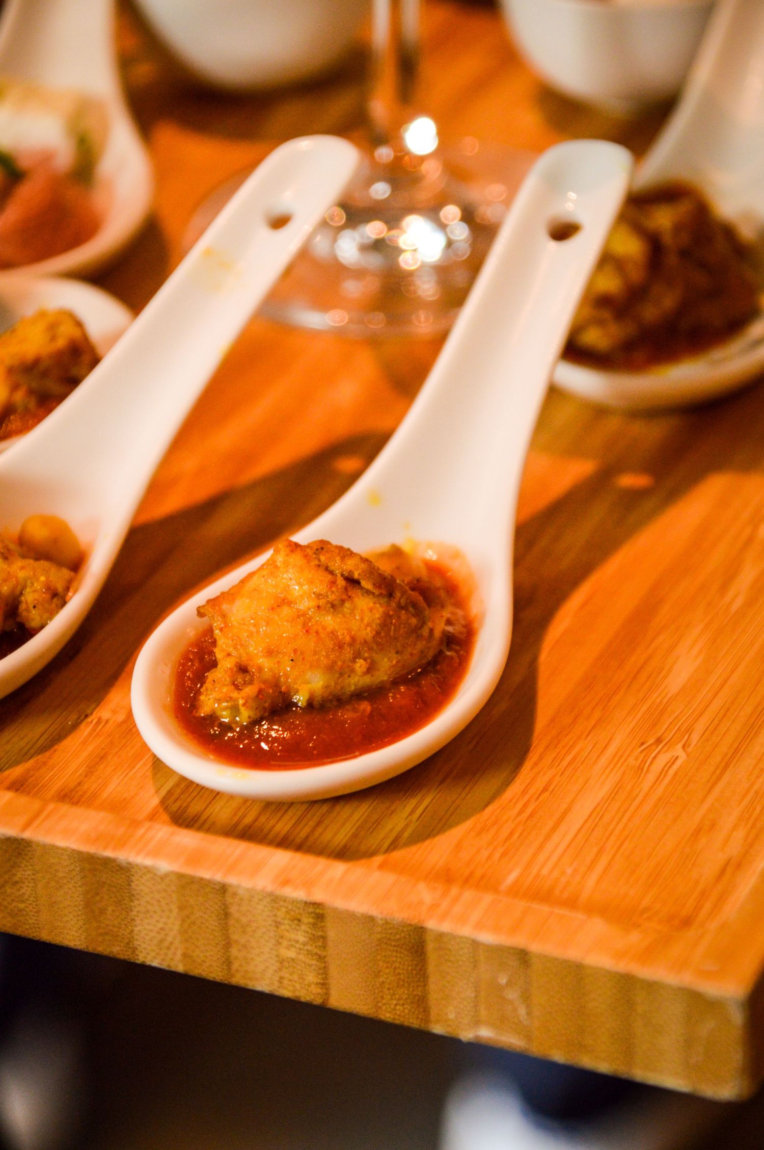 Appetizer spoon filled with a crispy fried morsel in red sauce on wooden serving board.