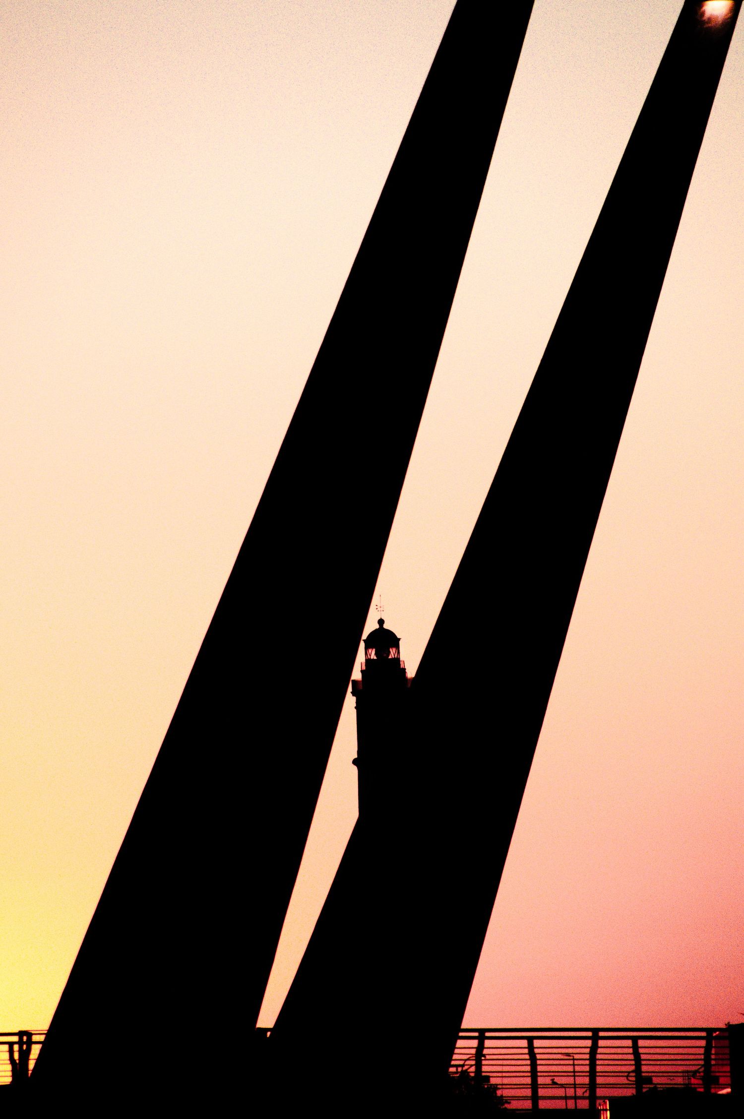 Silhouette of two tall bridge towers framing a lighthouse at sunset with warm orange and pink sky.