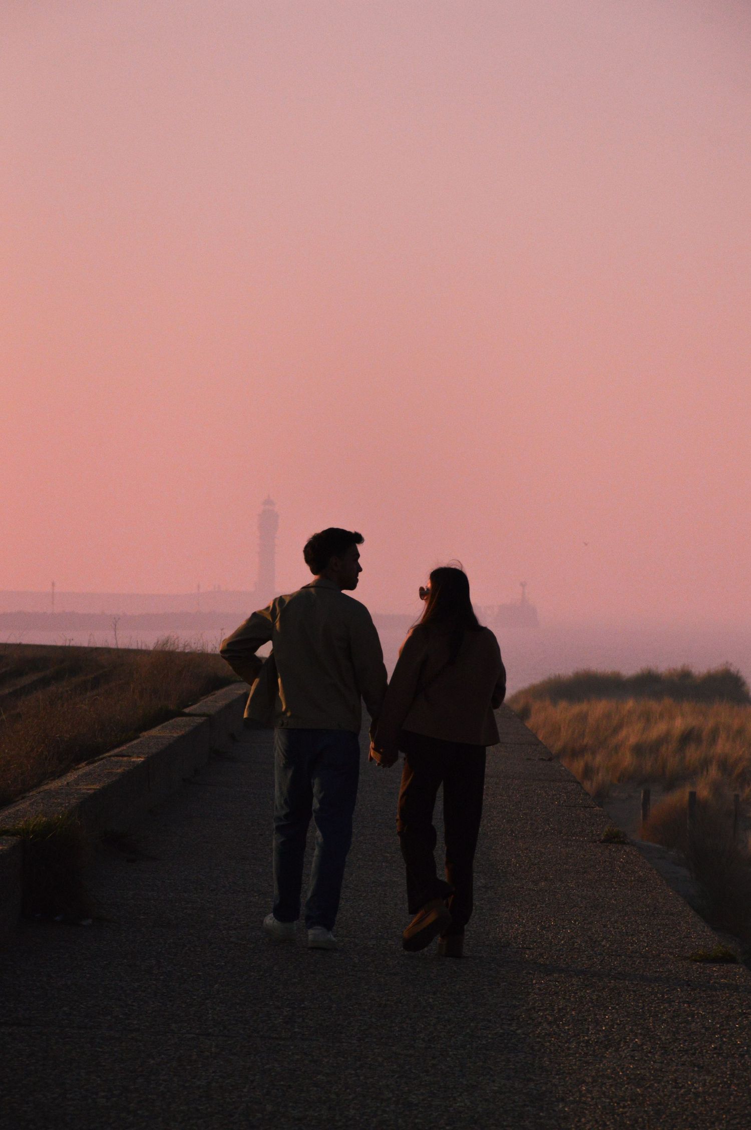 Deux silhouettes marchant sur un chemin au coucher du soleil, avec le phare du Risban à Dunkerque visible dans un arrière-plan brumeux.