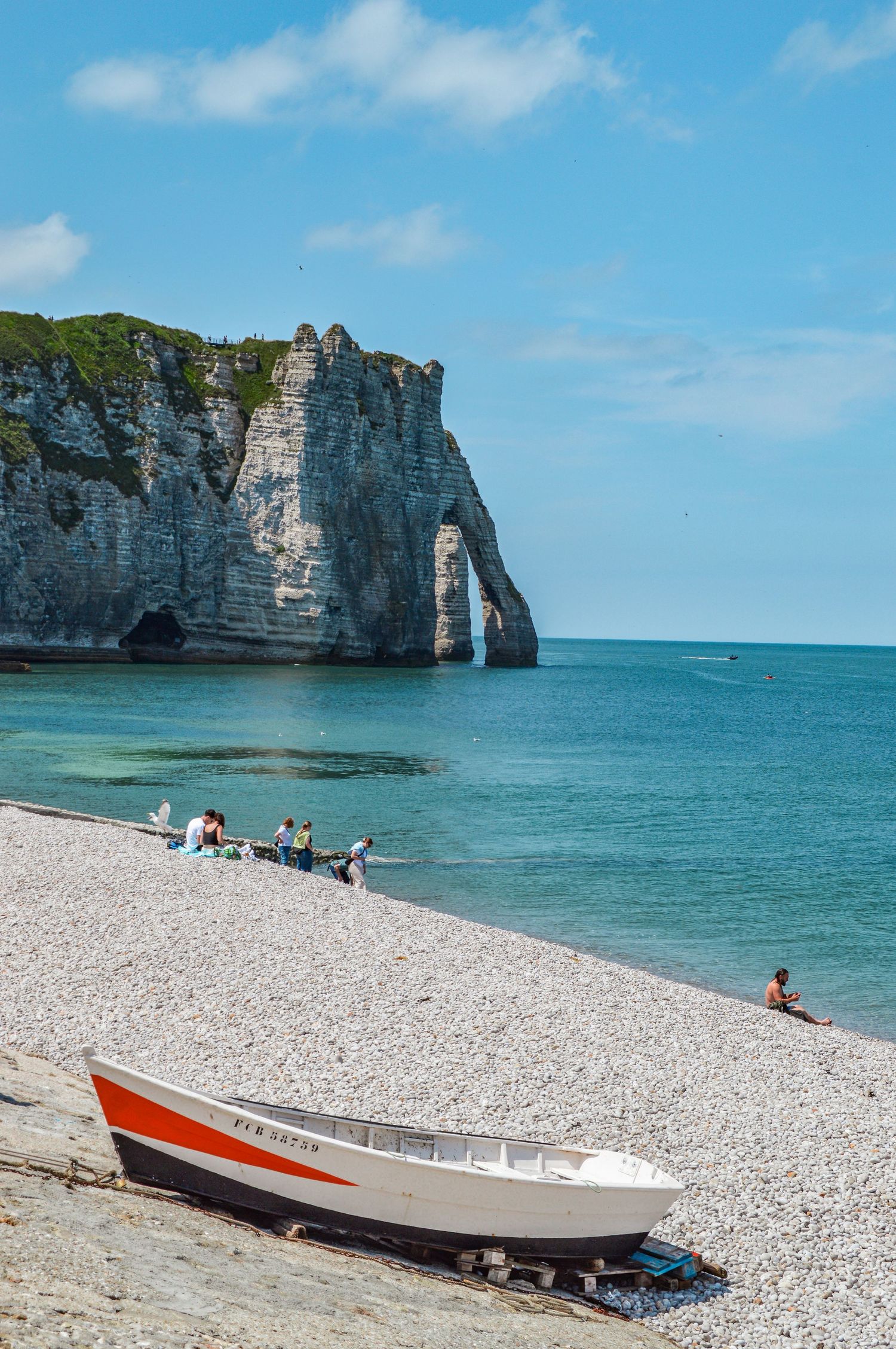 A red and white boat rests on the pebble beach with Etretat's famous cliff arch in the background.
