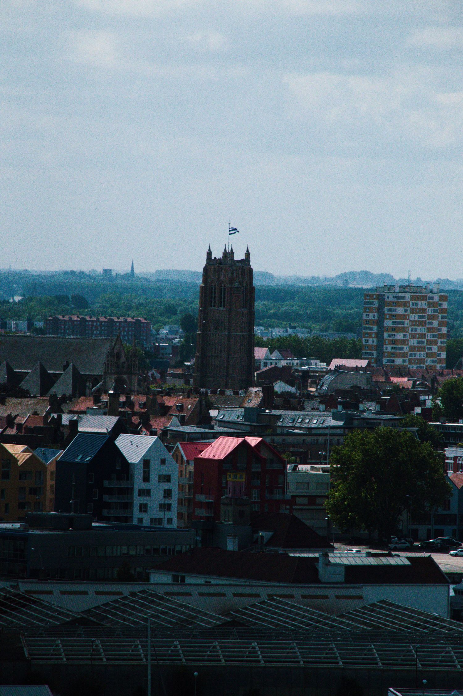 Aerial cityscape view featuring a historic Gothic church tower with flag, colorful rooftops, and modern buildings under cloudy sky.