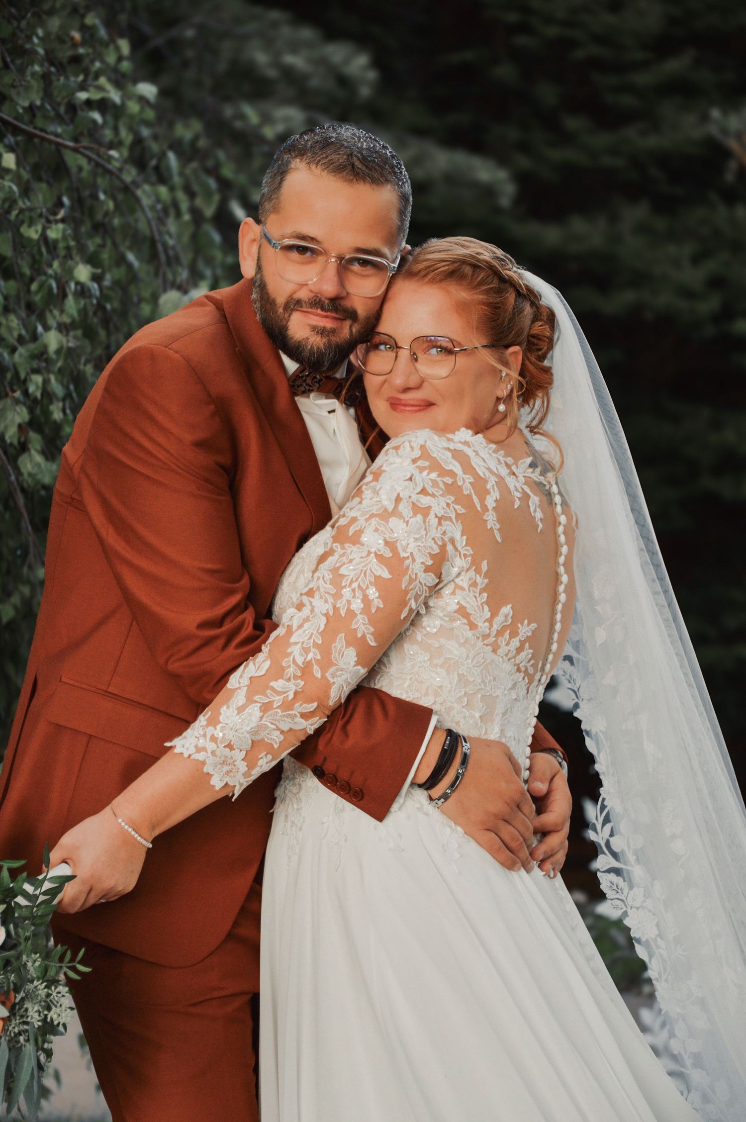 Romantic outdoor wedding photos featuring a couple in rust-colored suit and white lace dress against natural greenery.