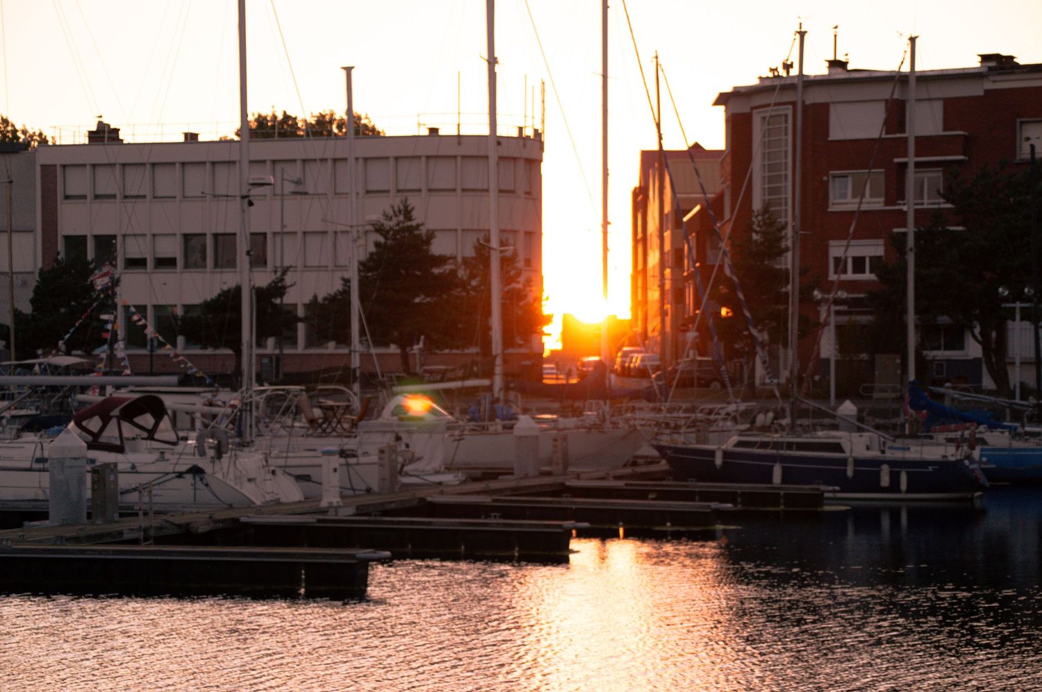 Sailboats docked in a marina at golden sunset with city buildings silhouetted against warm orange sky.