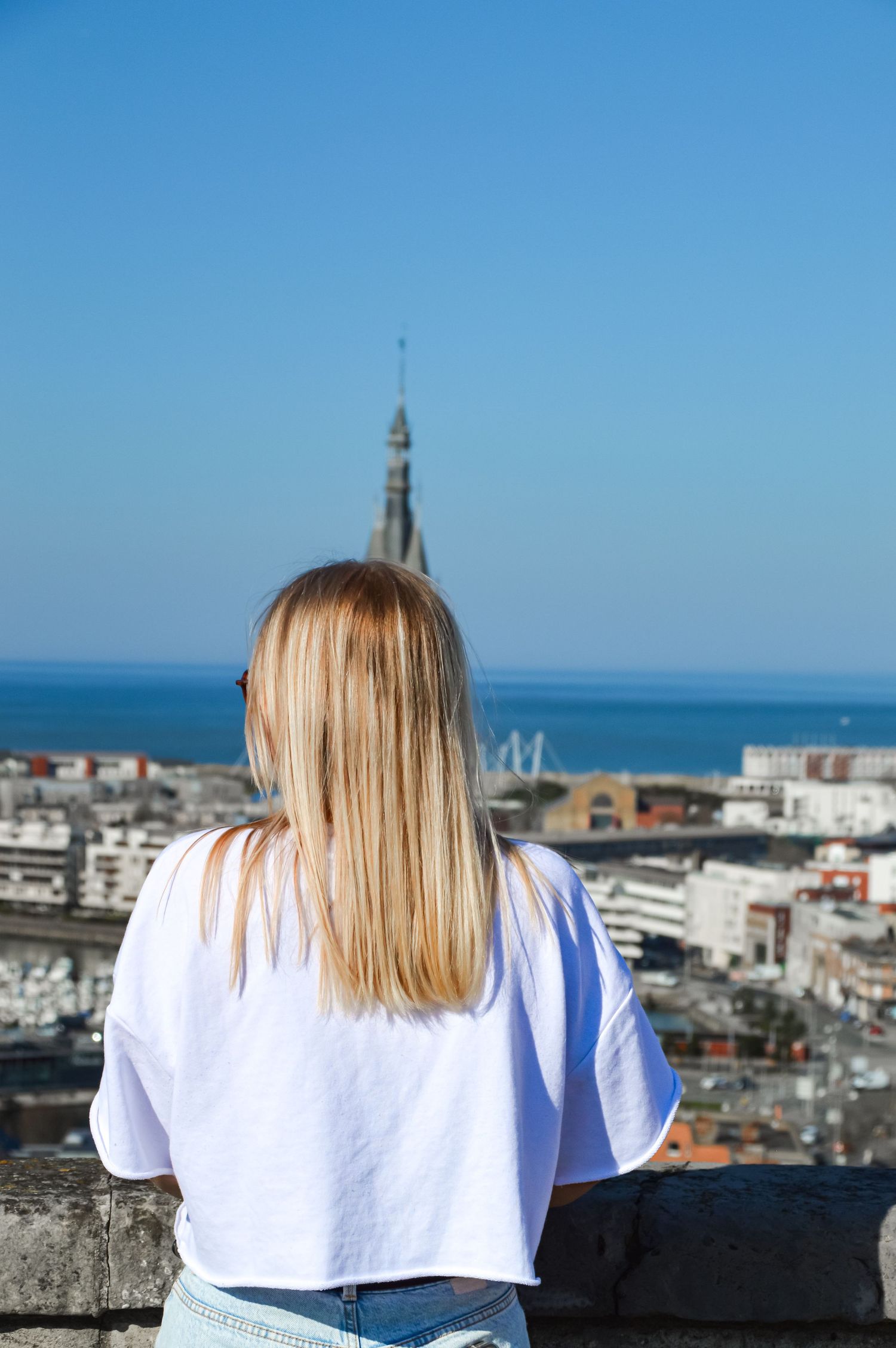 Scenic coastal city view with blue sky and person standing at stone wall overlook.