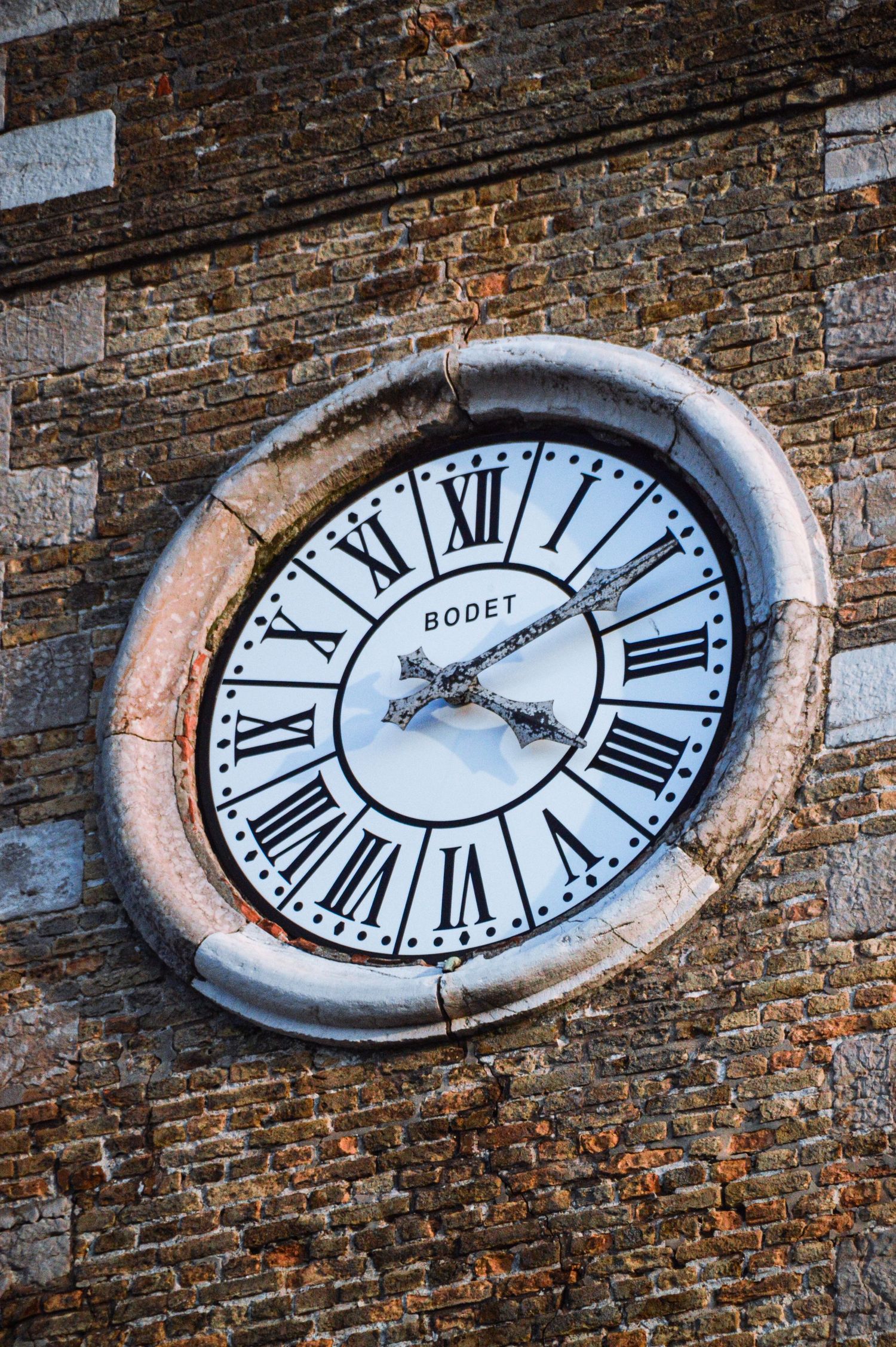 Vintage oval clock with Roman numerals mounted on weathered brick wall.