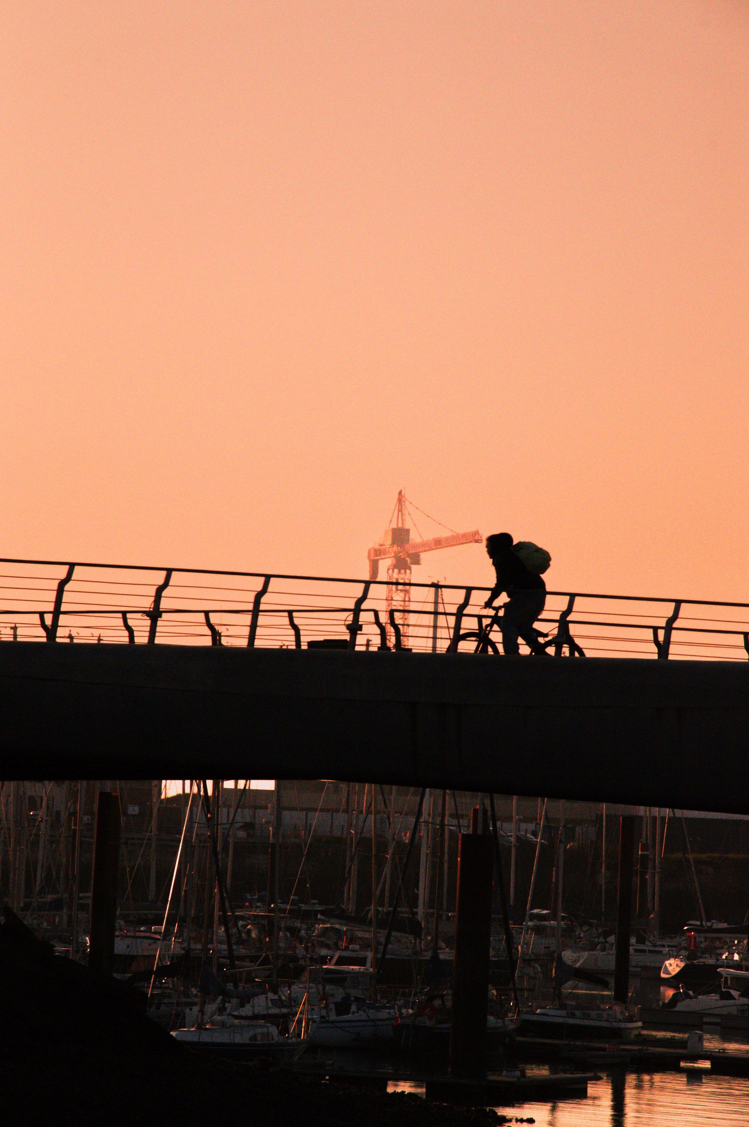 Silhouettes of people on a bridge at sunset with boats in the marina below creating a dramatic orange sky backdrop.