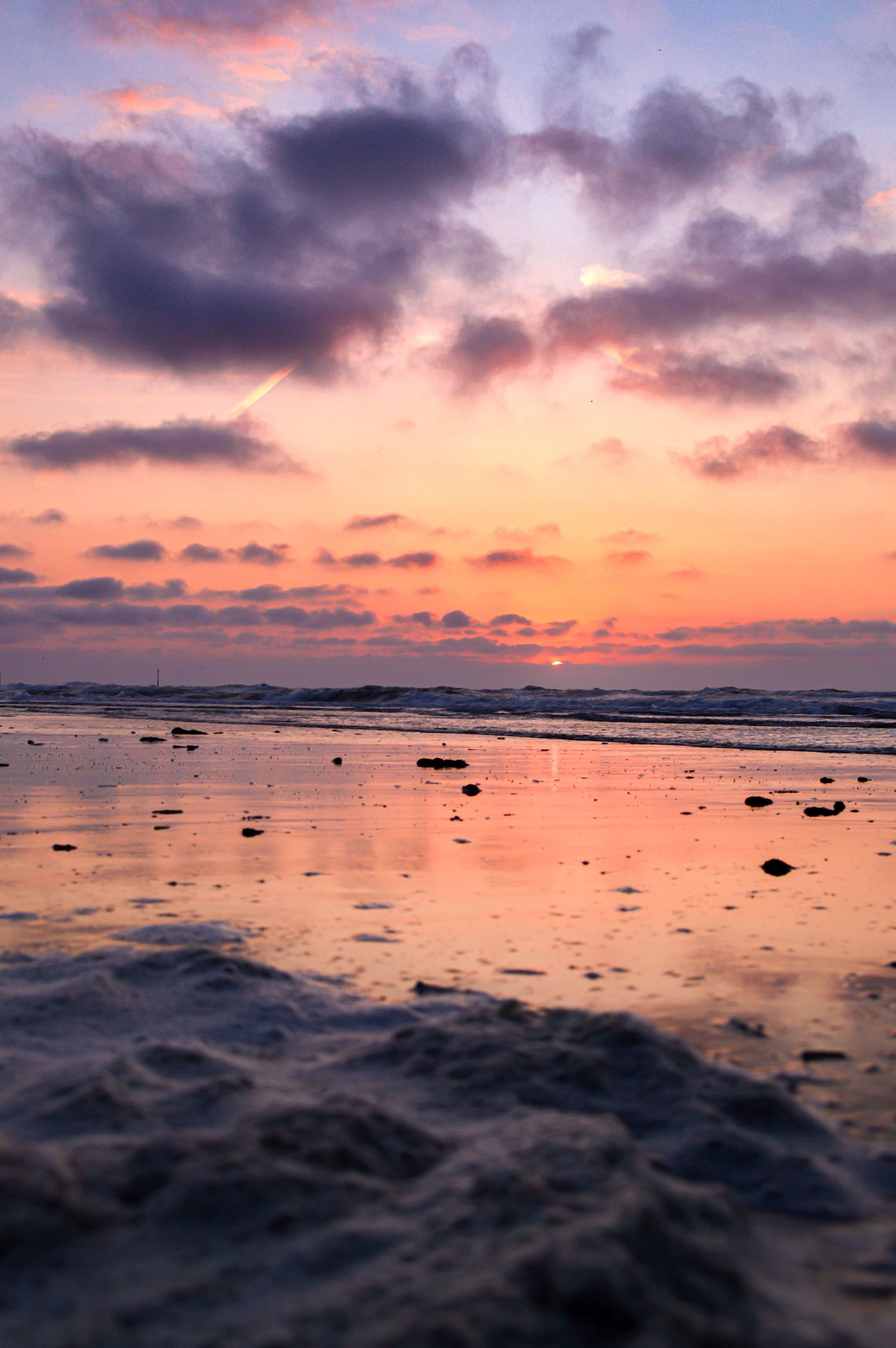Dramatic purple and orange sunset clouds reflect on wet beach sand with scattered rocks.