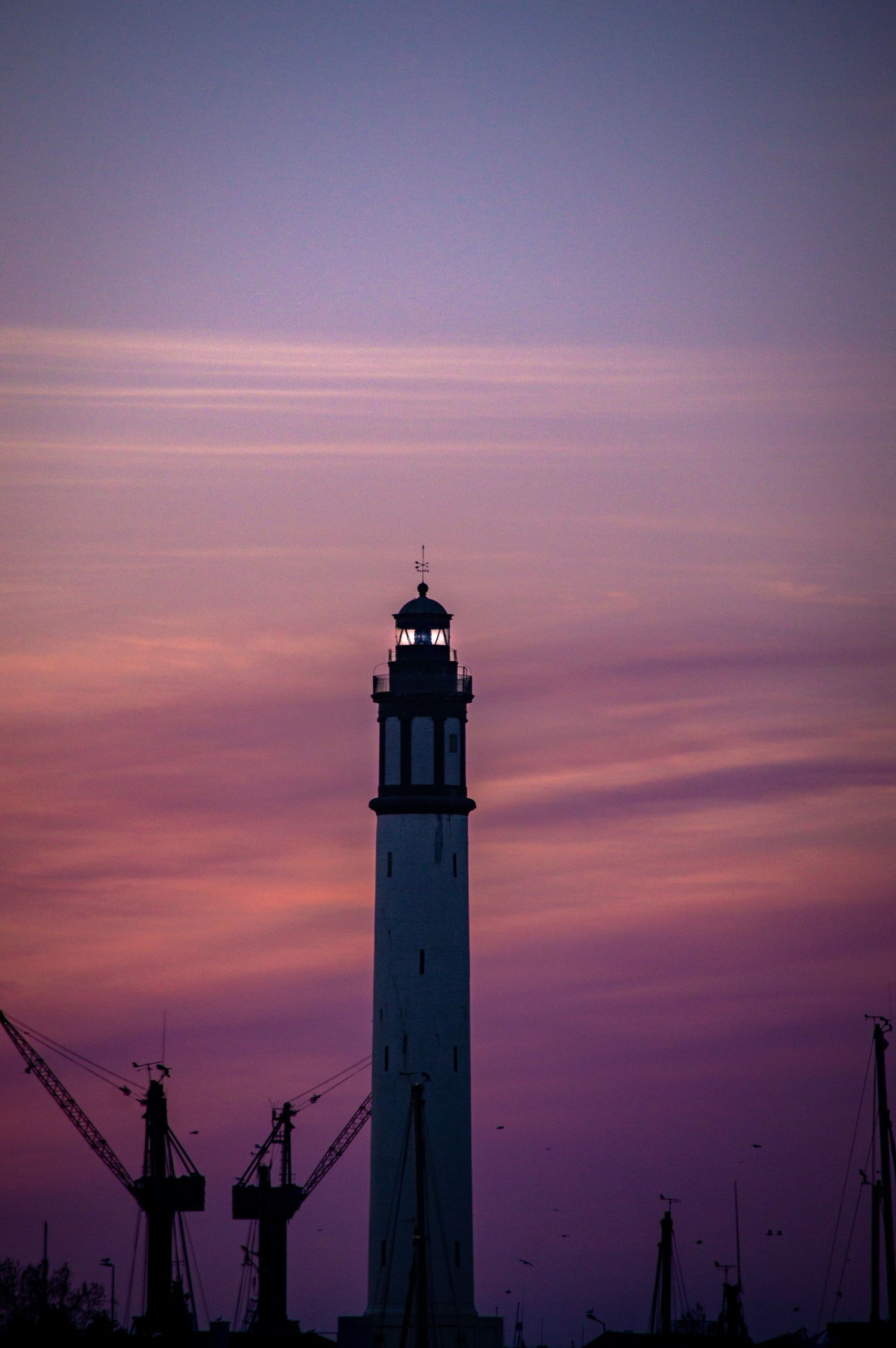 Silhouette of a lighthouse against a vibrant purple and pink sunset sky with industrial cranes visible in the distance.