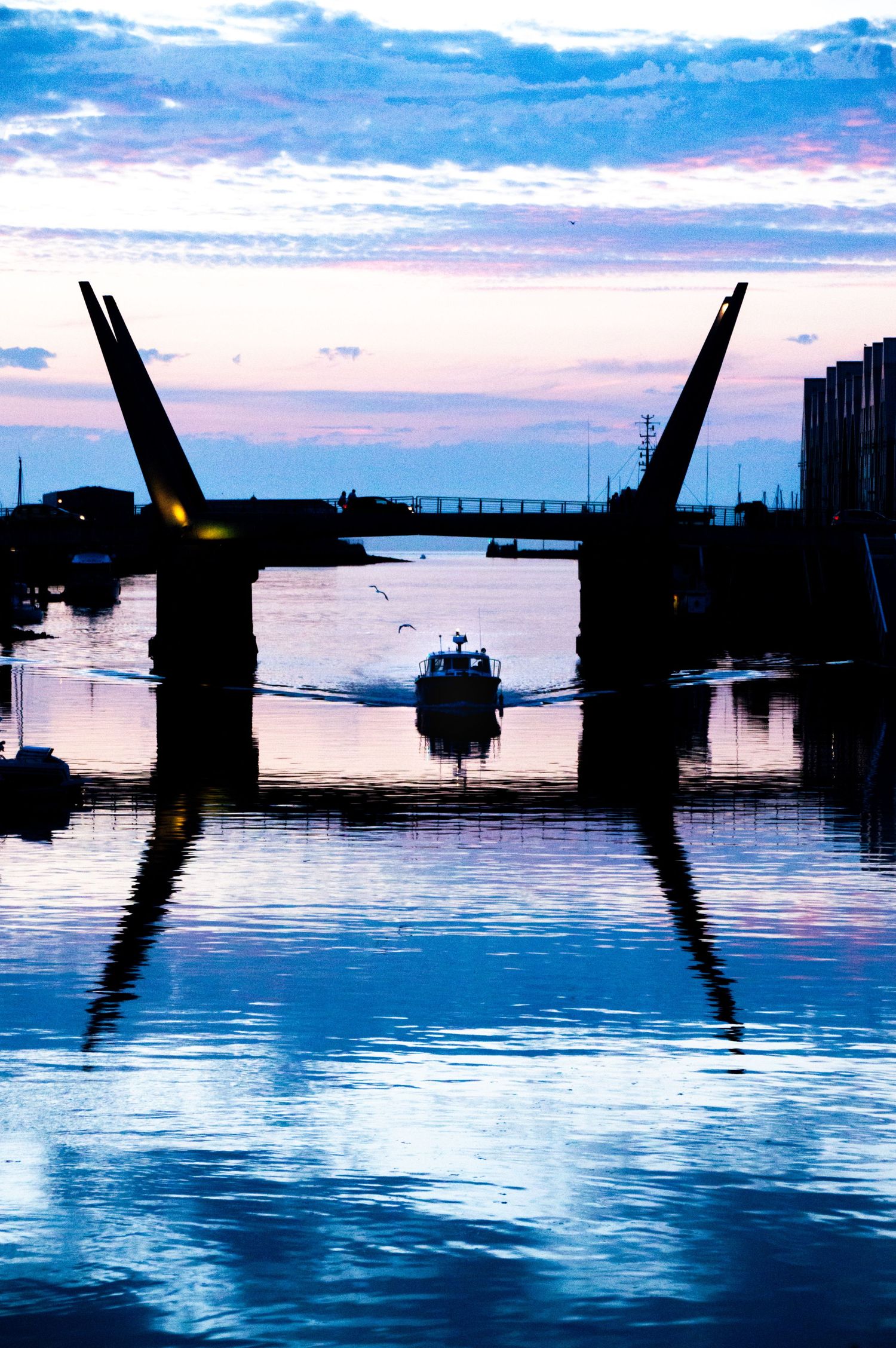 A silhouetted drawbridge opens at dusk as a boat passes through calm blue waters reflecting a purple and pink sunset sky.