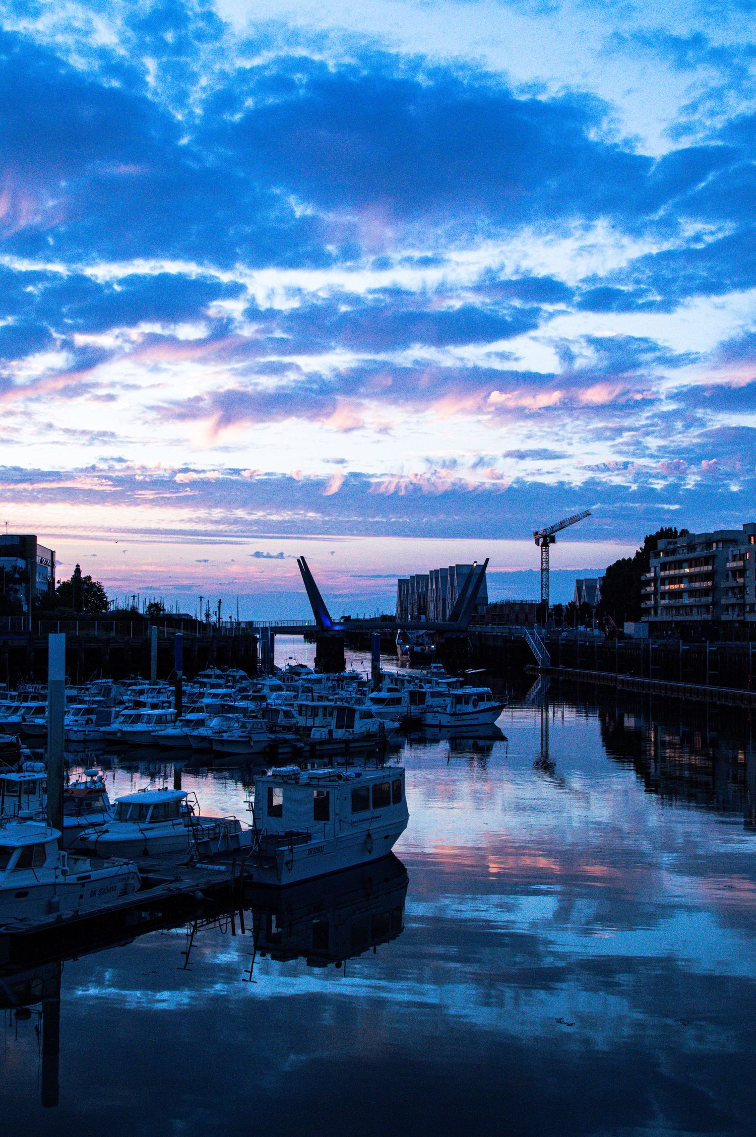 Blue hour marina with boats reflecting in calm water, dramatic cloudy sky at dusk, urban waterfront with crane silhouette.