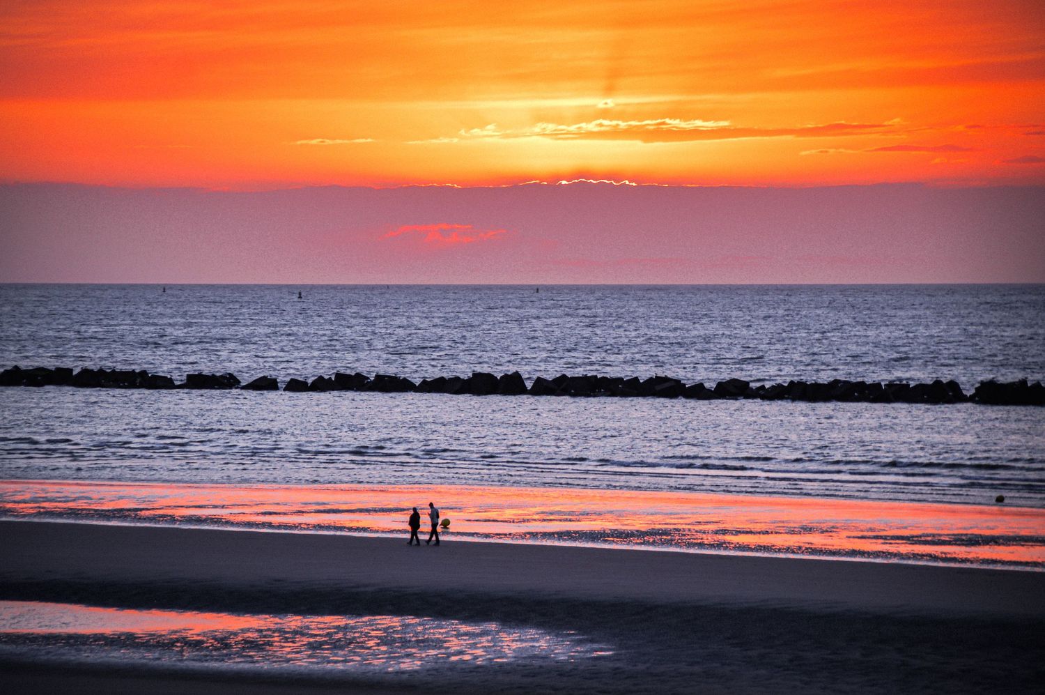 Dramatic orange and pink sunset over ocean horizon with silhouettes on beach.