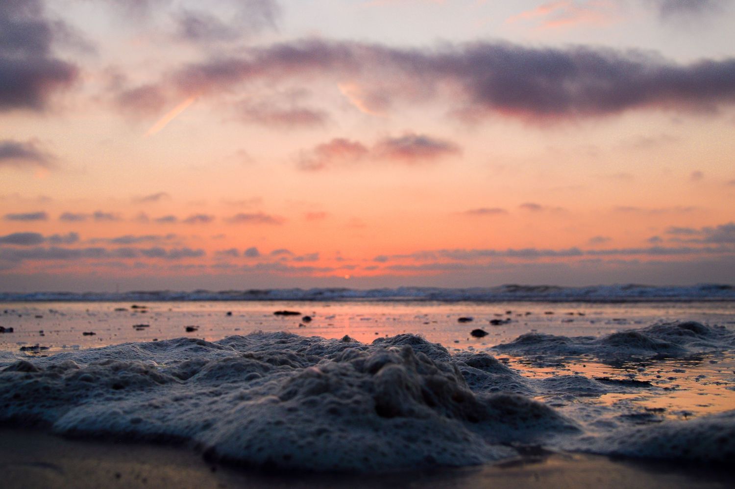 Dramatic ocean sunset with orange sky reflecting on wet sand and scattered rocks.