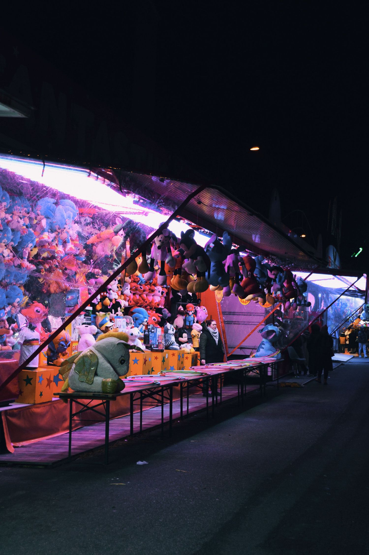 Brightly lit carnival gaming stalls stretching into the distance at night.