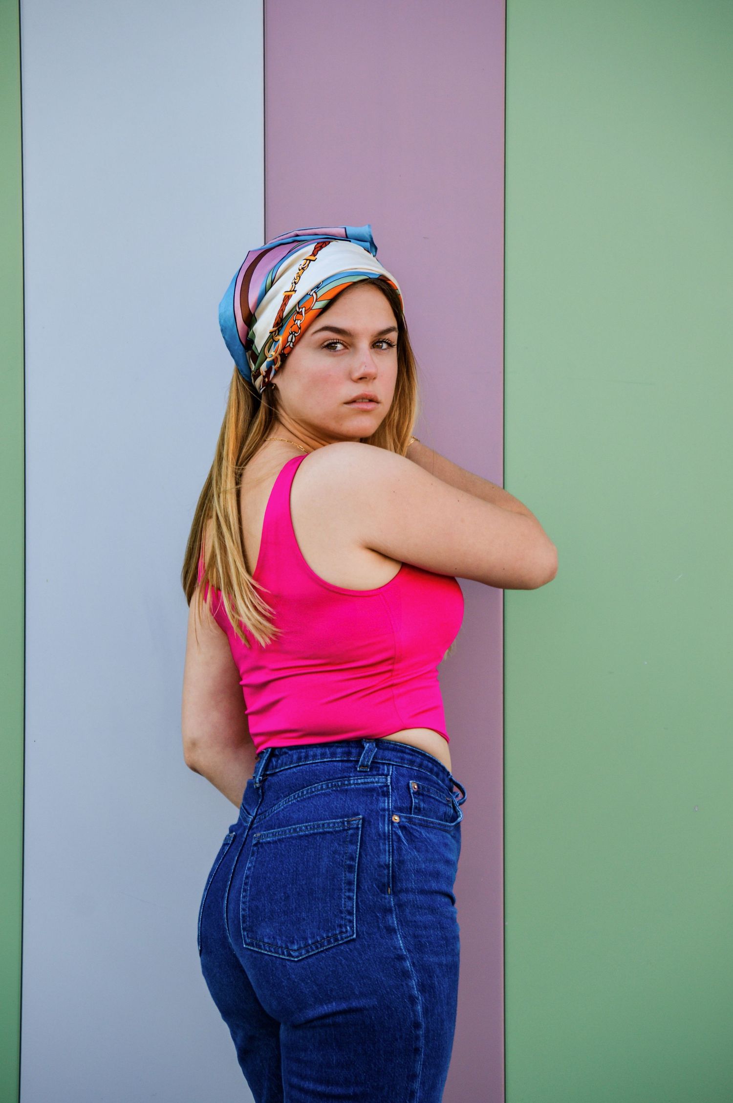 Model in pink top and jeans poses against colorful striped wall backdrop.