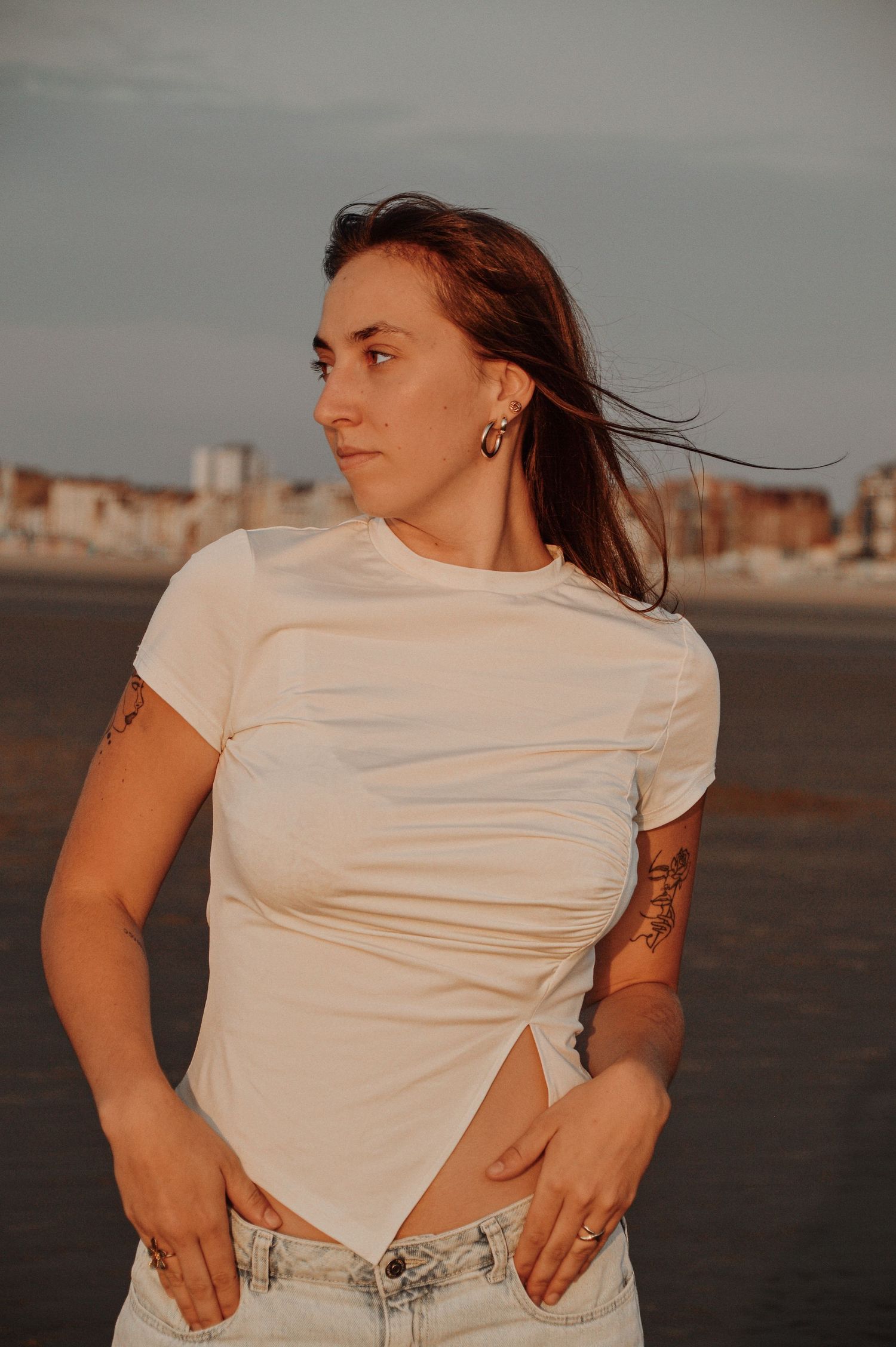 Woman in white t-shirt and jeans poses against urban backdrop at sunset.