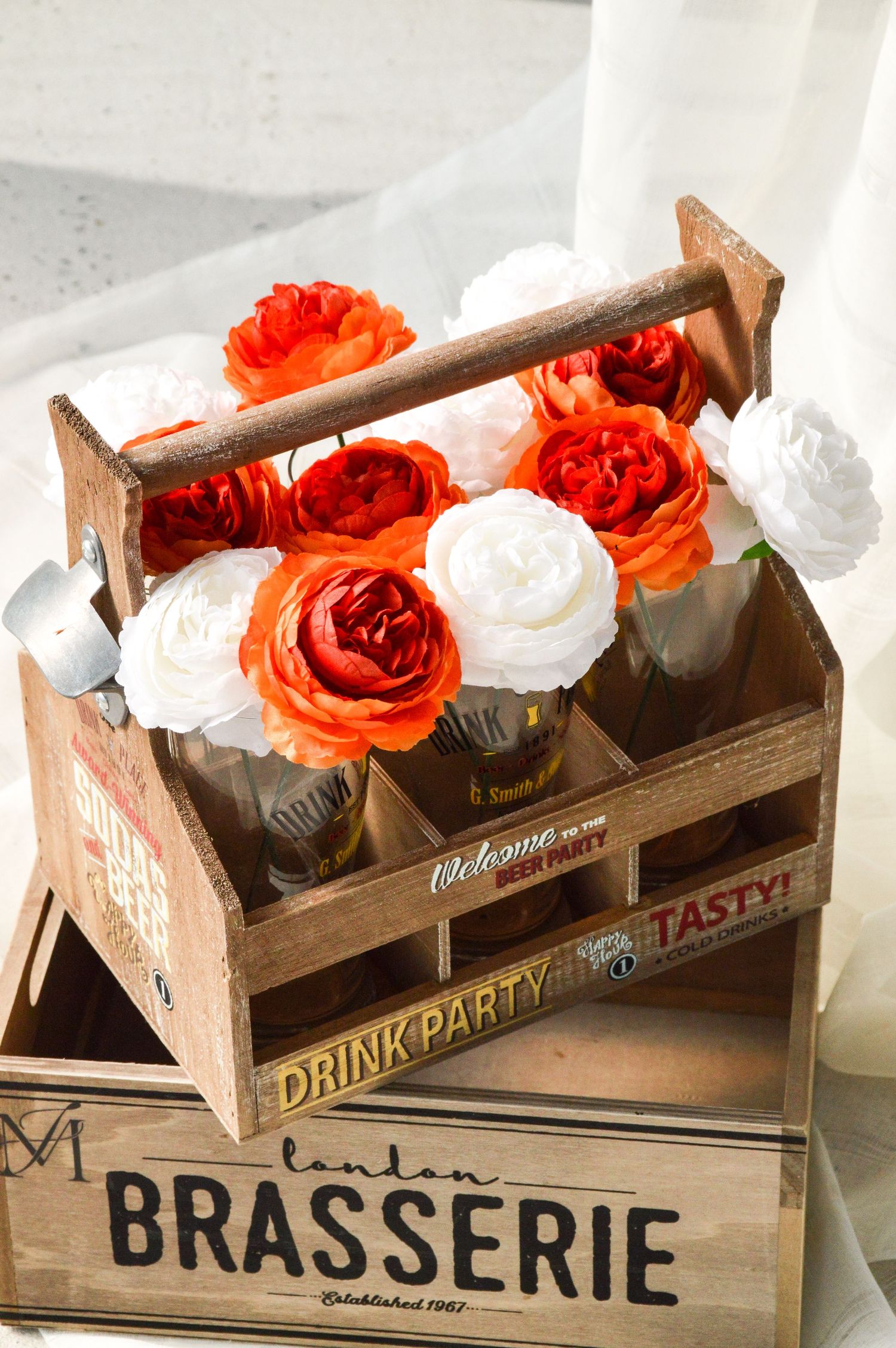 Rustic wooden crate labeled 'Brasserie' holds orange roses and white flowers in glass vases.