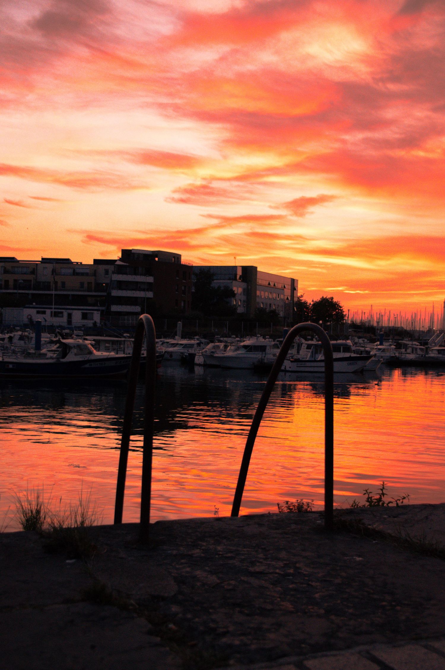 Vibrant red and orange sunset over a marina with silhouetted boats and buildings reflecting on calm water.