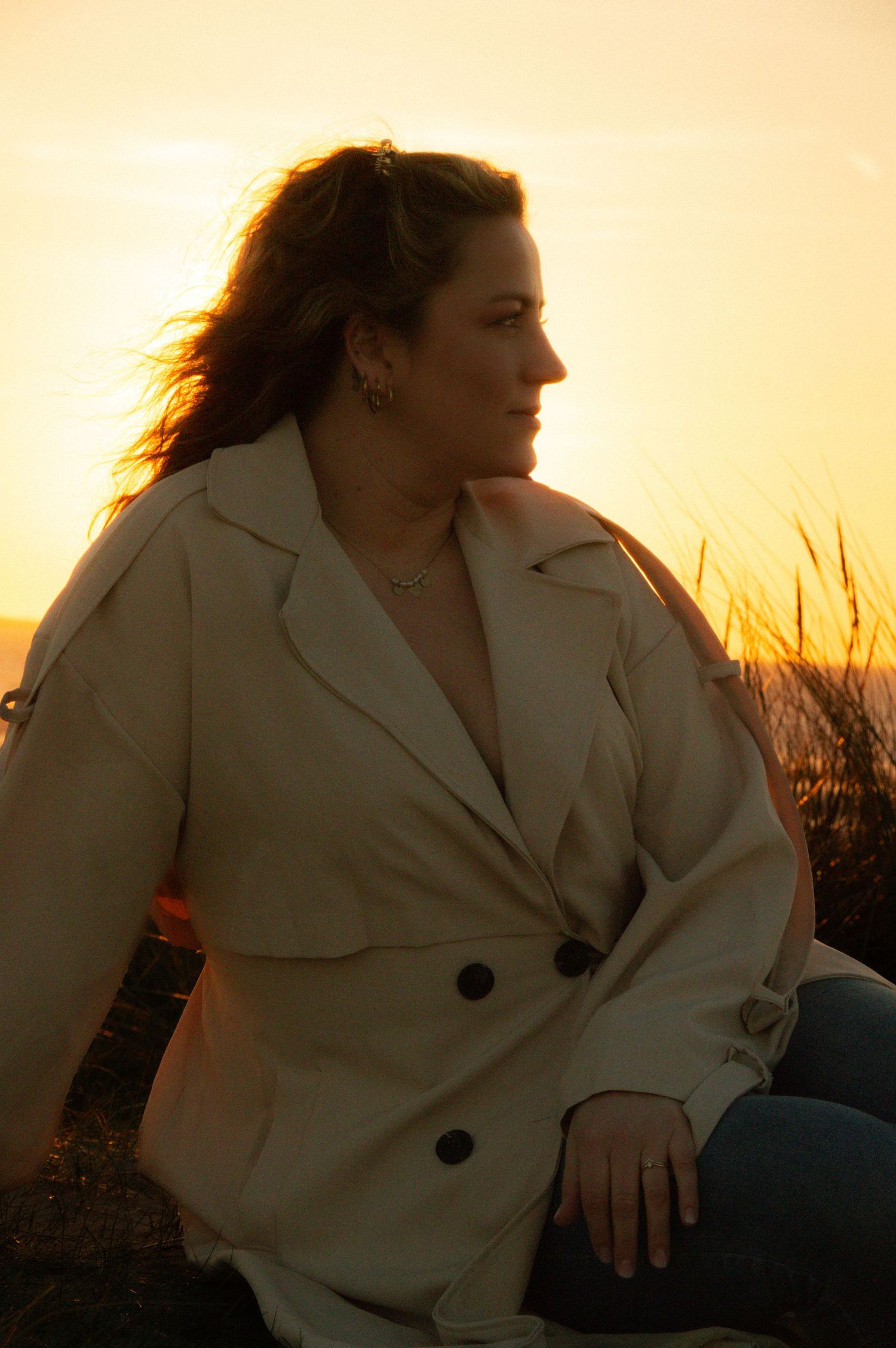 Woman in beige trench coat sitting outdoors at golden sunset with wind in her hair.