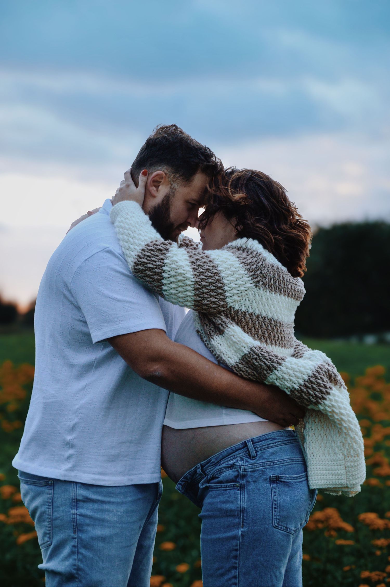 A couple embraces in a field of flowers during sunset wearing casual clothing.
