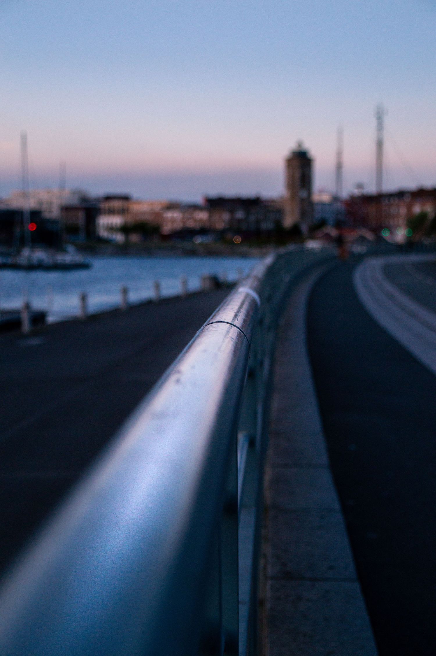 A shiny metal railing curves along a waterfront promenade at dusk with a city skyline softly blurred in the background.