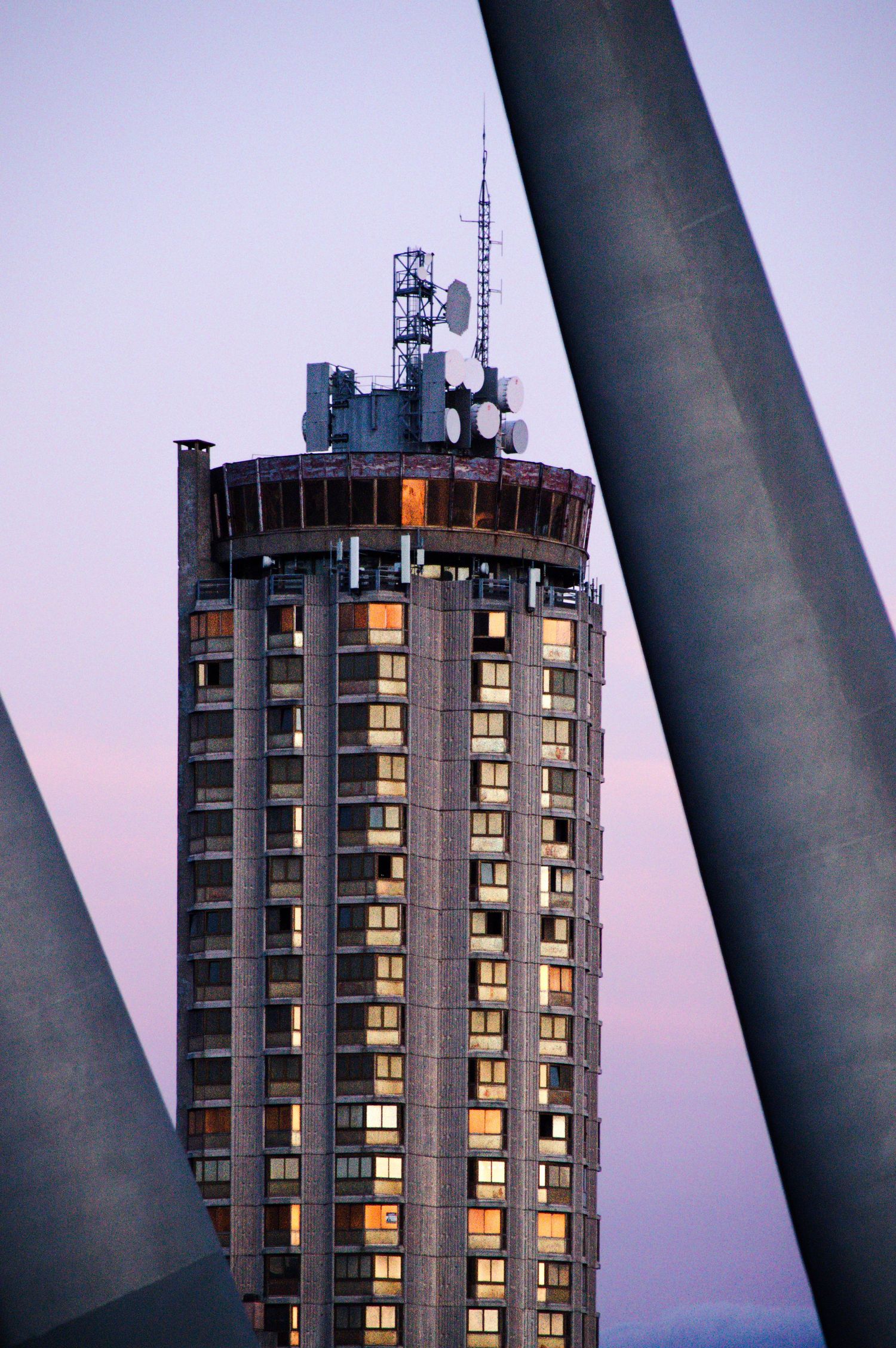 Tall cylindrical skyscraper with communication antennas on top, framed by concrete pillars at dusk with purple sky.