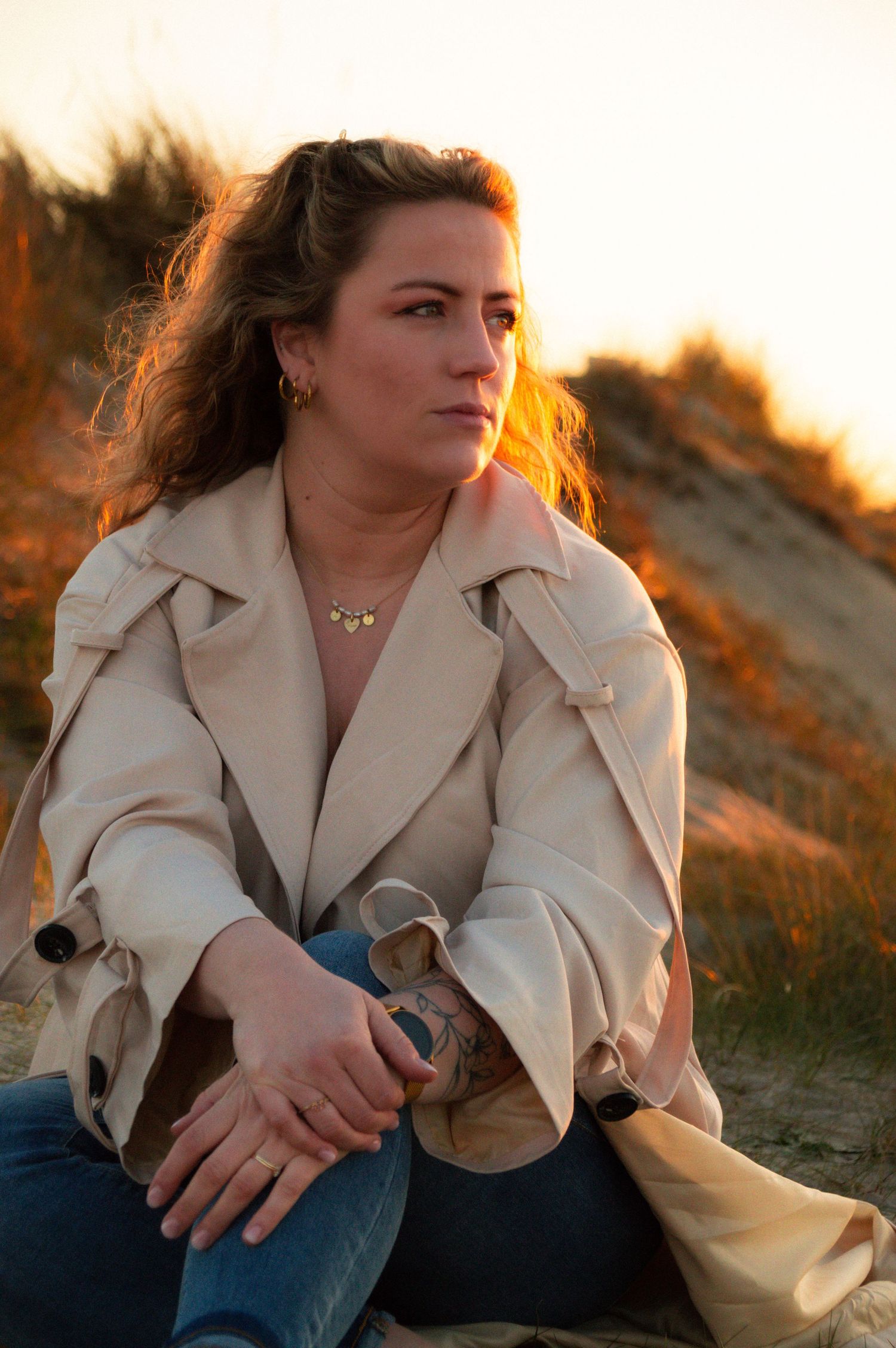 Woman wearing beige coat and gold necklace sitting on sandy dunes during warm sunset.