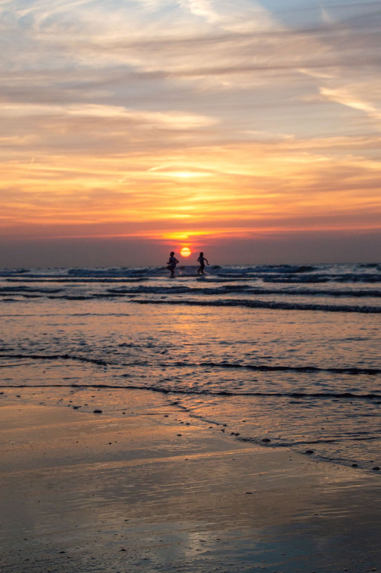 Two children silhouetted against a vibrant orange and pink sunset over ocean waves on a sandy beach.
