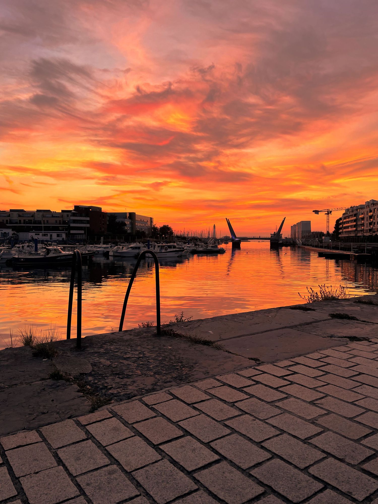 Stunning orange and red sunset over a calm harbor with boats, reflecting vibrant colors on the water beside a cobblestone walkway.