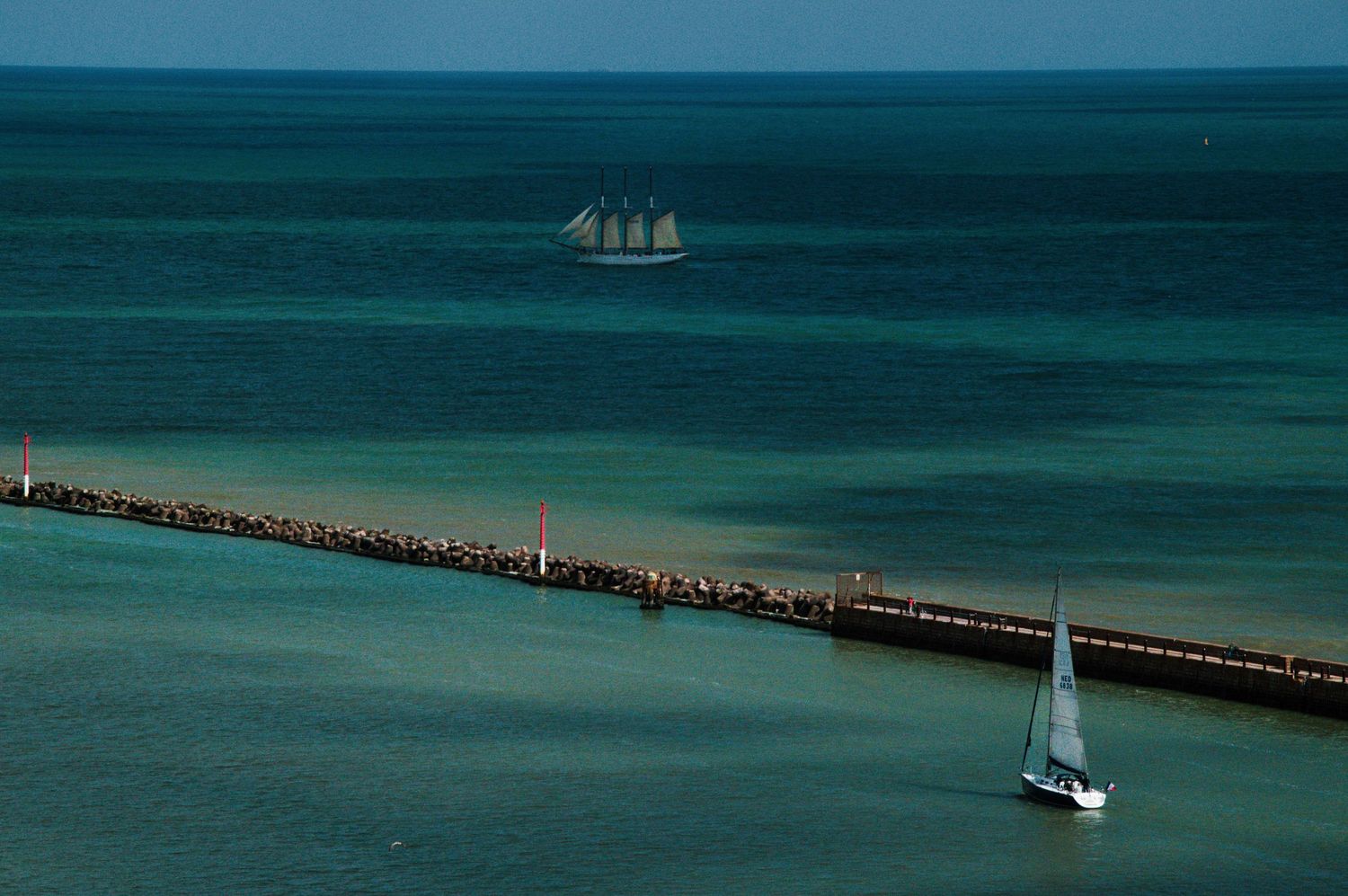 Aerial view of a sailboat near a stone pier with a tall ship sailing on turquoise ocean waters in the distance.