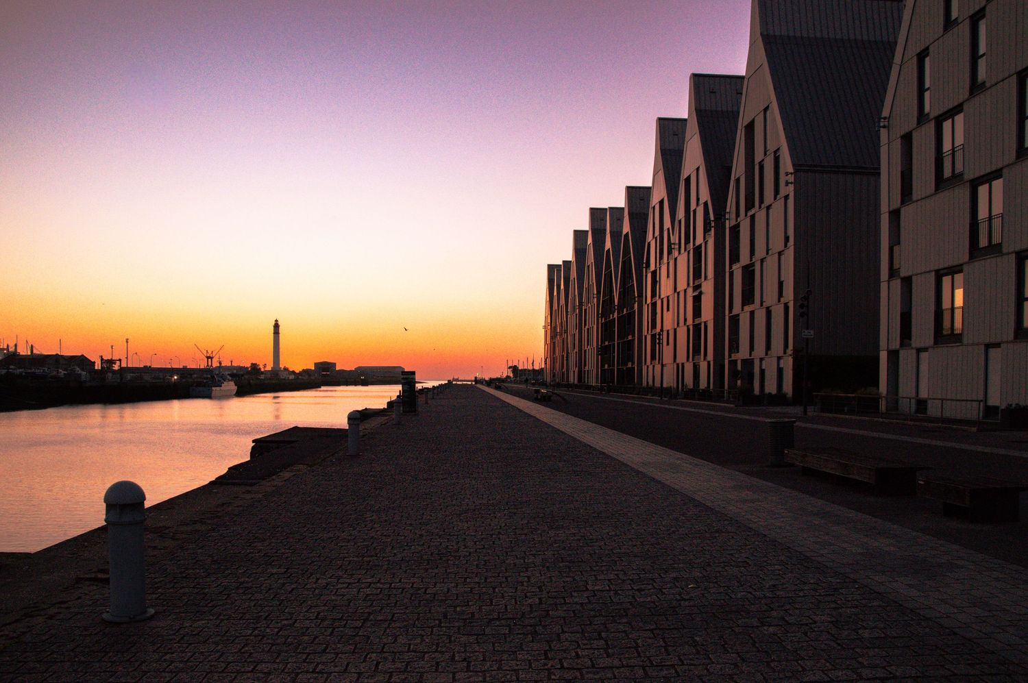 Sunset over a waterfront promenade with modern brick buildings and a colorful orange and purple sky reflected on calm water.