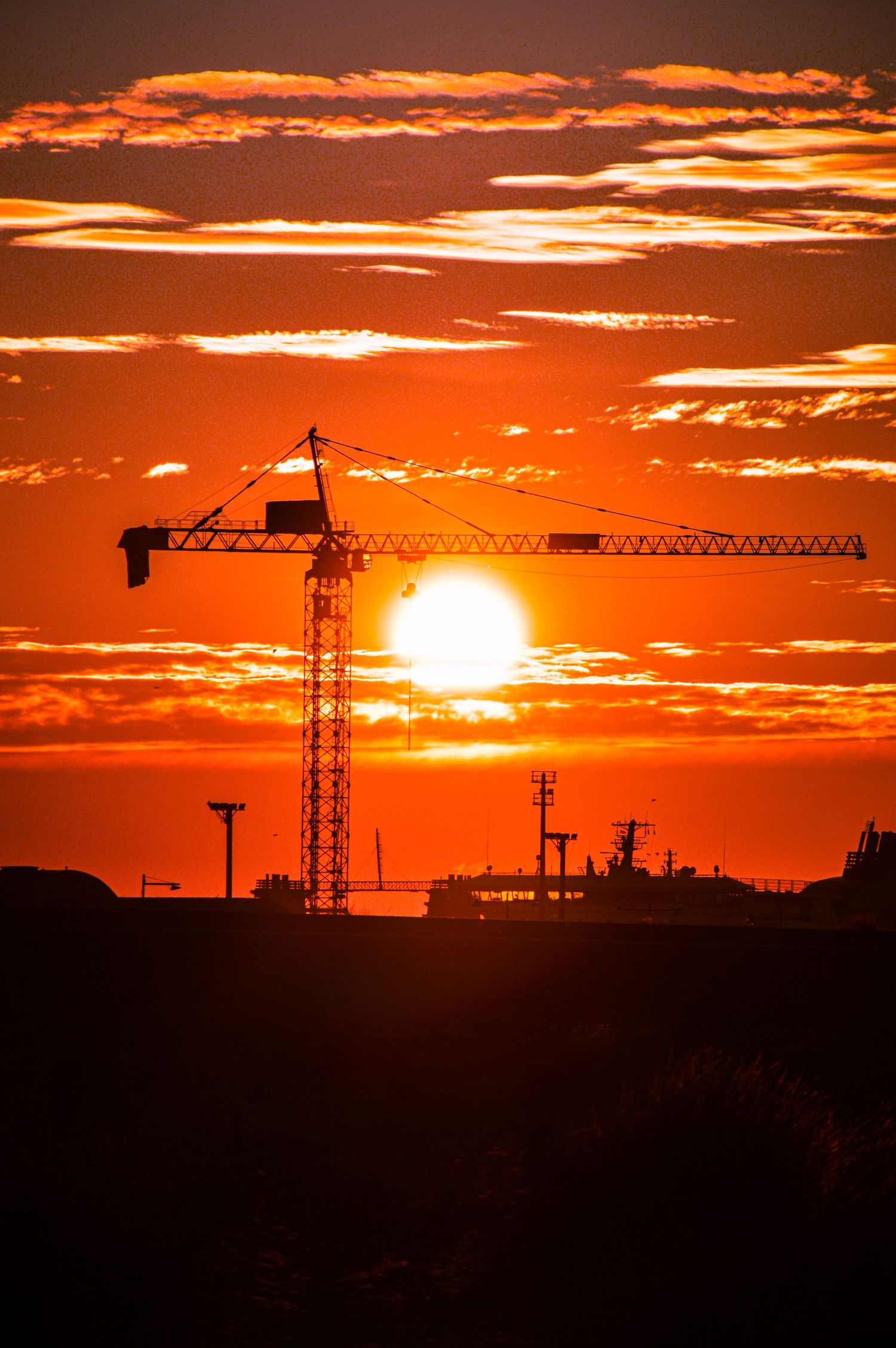 Construction cranes silhouetted against a dramatic orange sunset sky with glowing sun on the horizon.