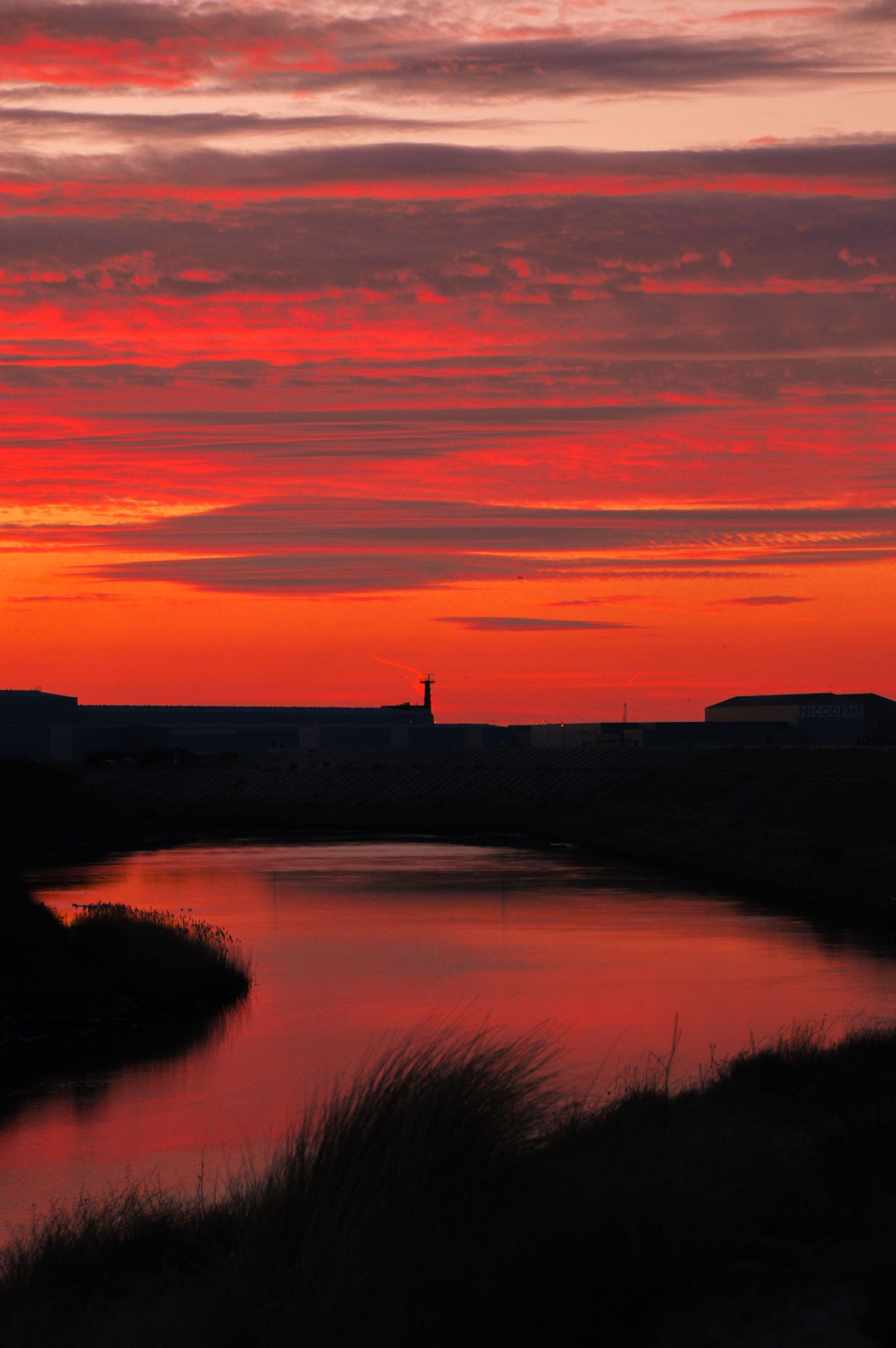 Silhouette of coastal landscape against brilliant red sunset sky with lighthouse in distance.