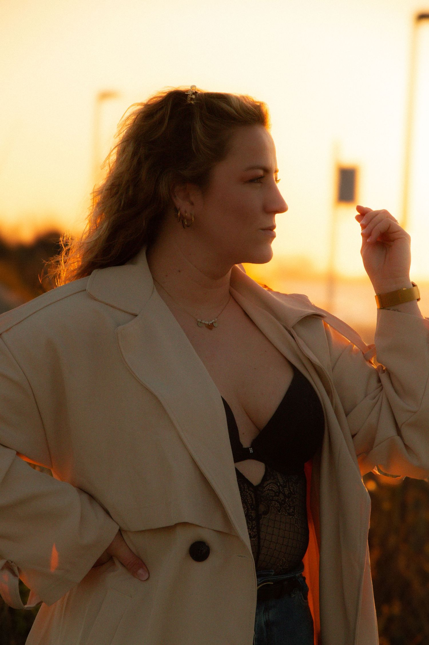 Woman in beige blazer and black lace top poses outdoors during golden hour sunset, wearing gold jewelry.