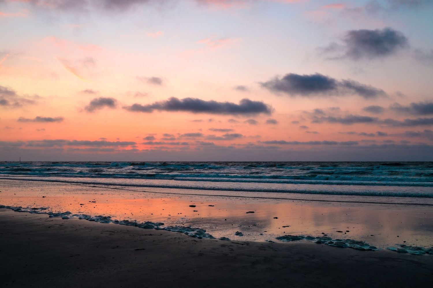 Colorful coastal sunset with waves rolling onto shore under pink and orange clouds.