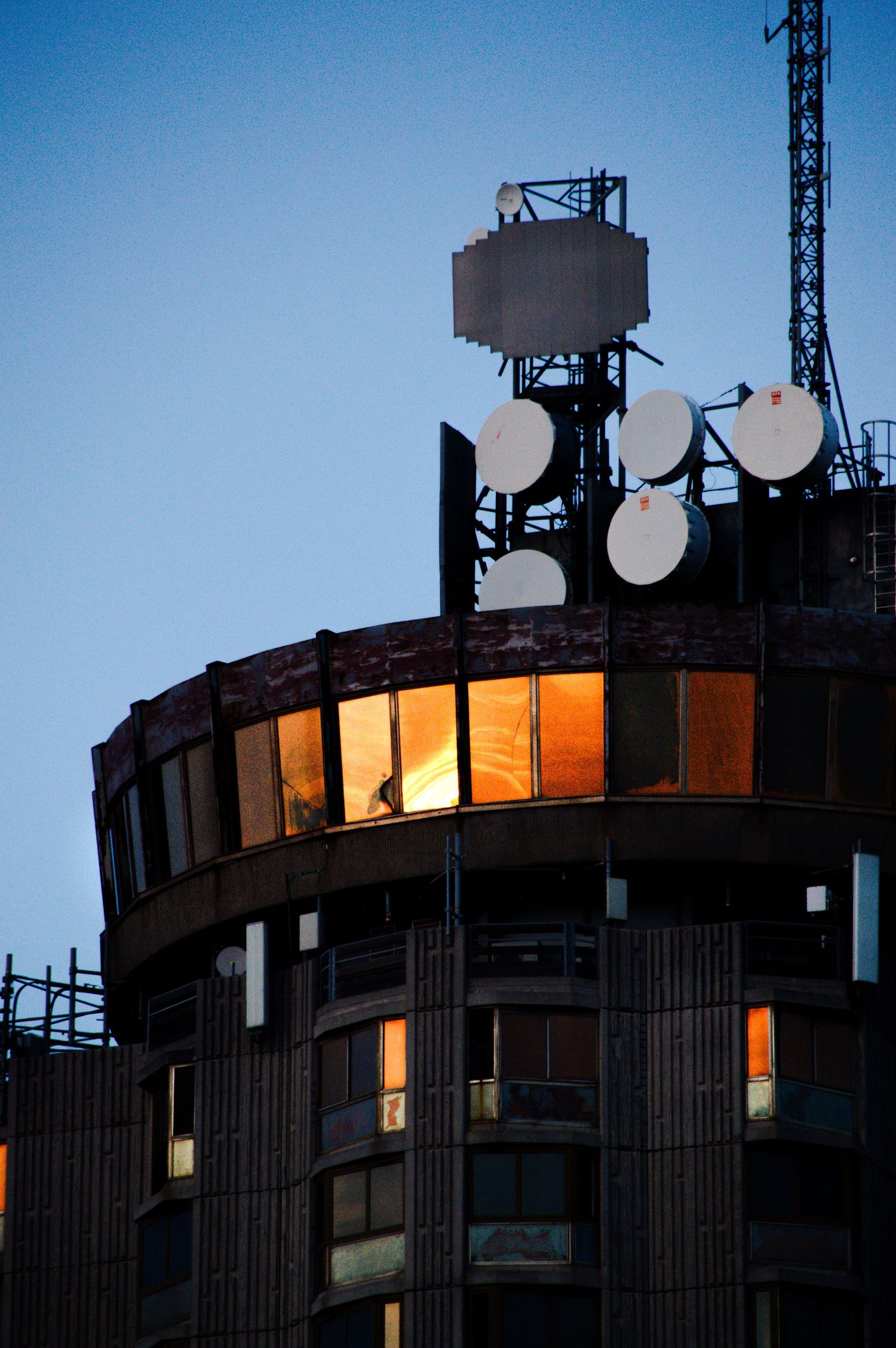 Circular urban building at dusk with illuminated windows and multiple satellite dishes and communication towers on rooftop.