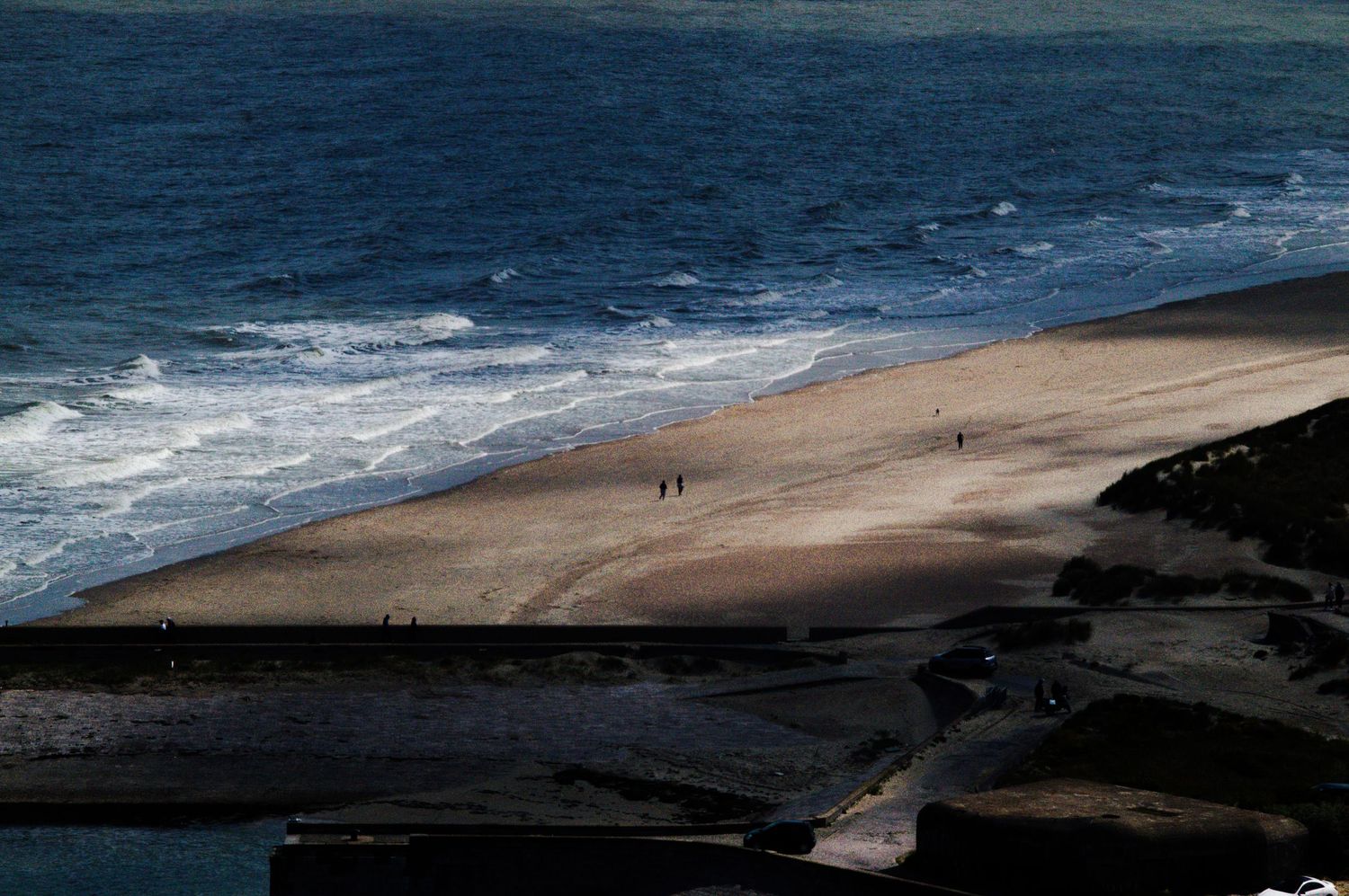 Aerial view of a sandy beach with ocean waves and people walking along the shoreline on a sunny day.