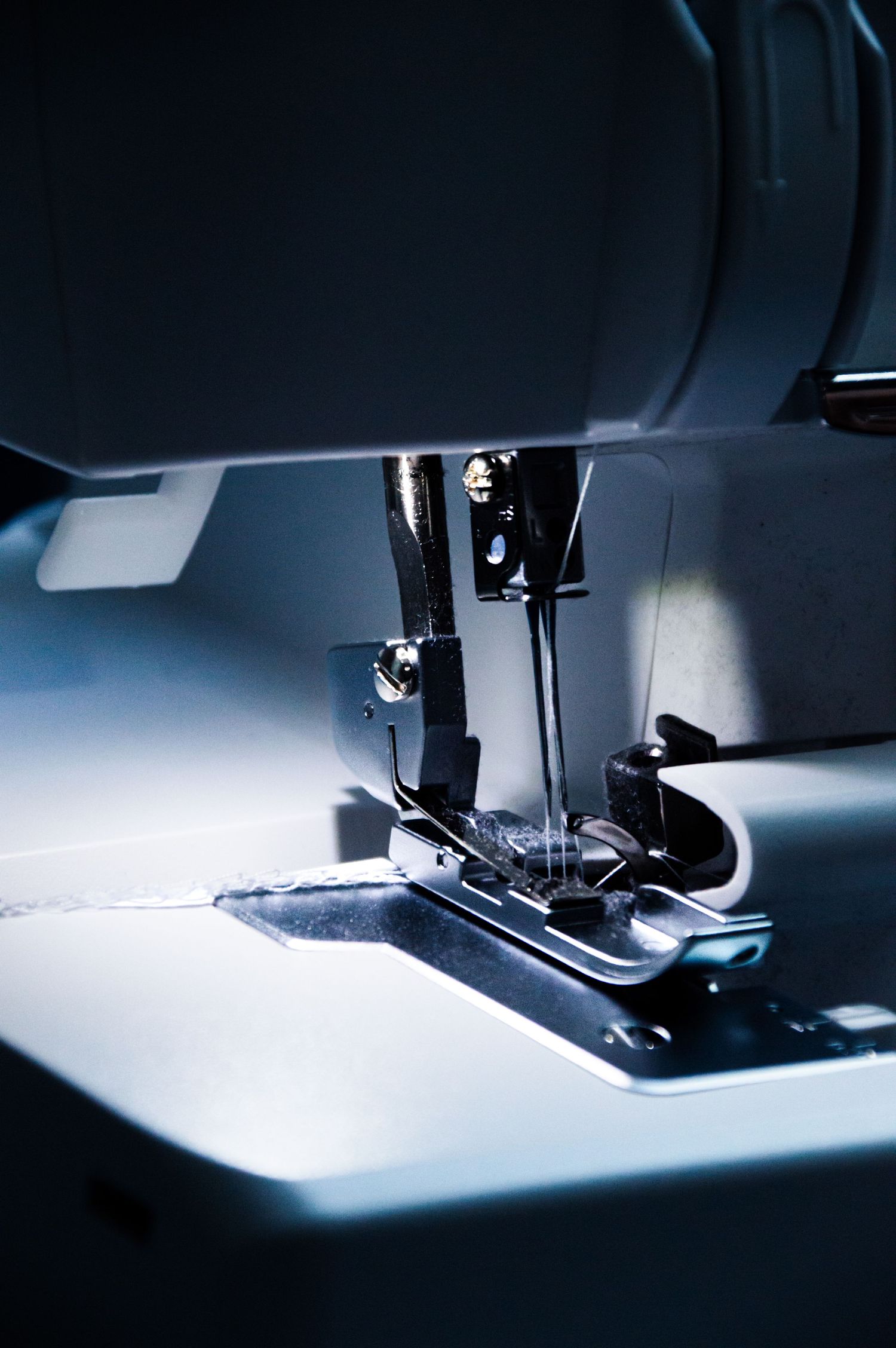 Close-up of a sewing machine needle and presser foot illuminated by light while stitching gray fabric.