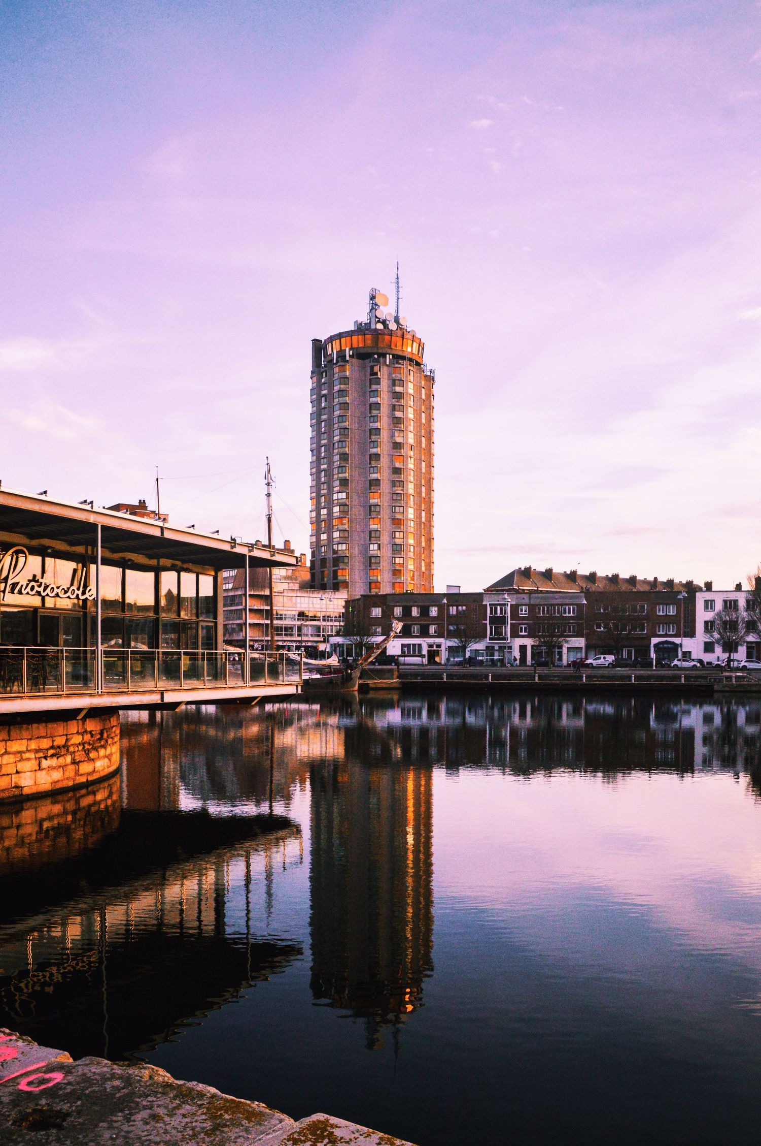 Waterfront cityscape with modern buildings reflecting in calm harbor at sunset.