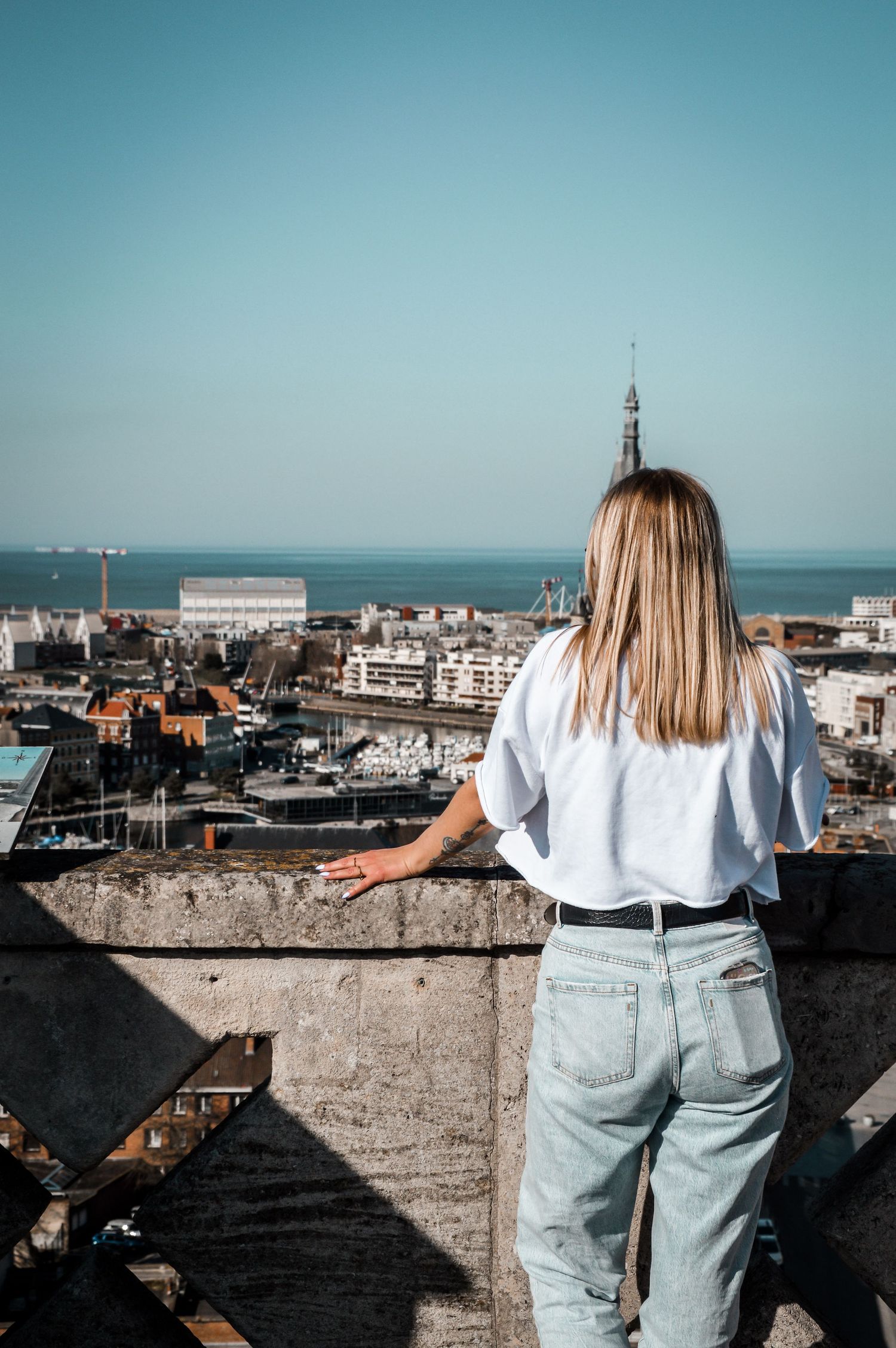 Person in light denim jeans and white shirt looks out over coastal cityscape from elevated viewpoint.