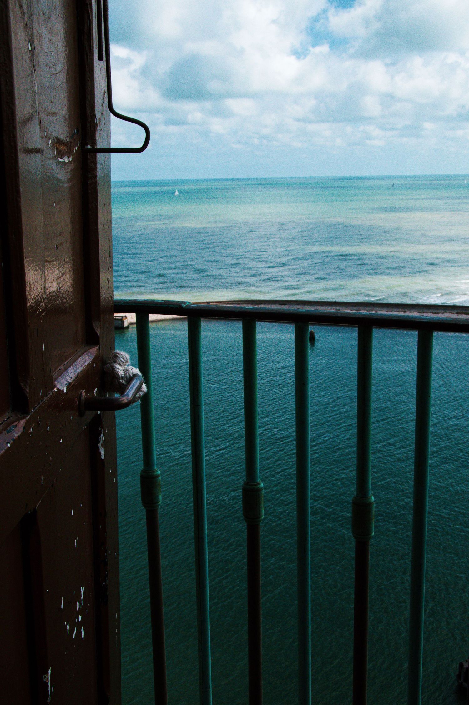 A metal railing balcony overlooks a stunning turquoise ocean under a partly cloudy sky, viewed from a weathered doorway.