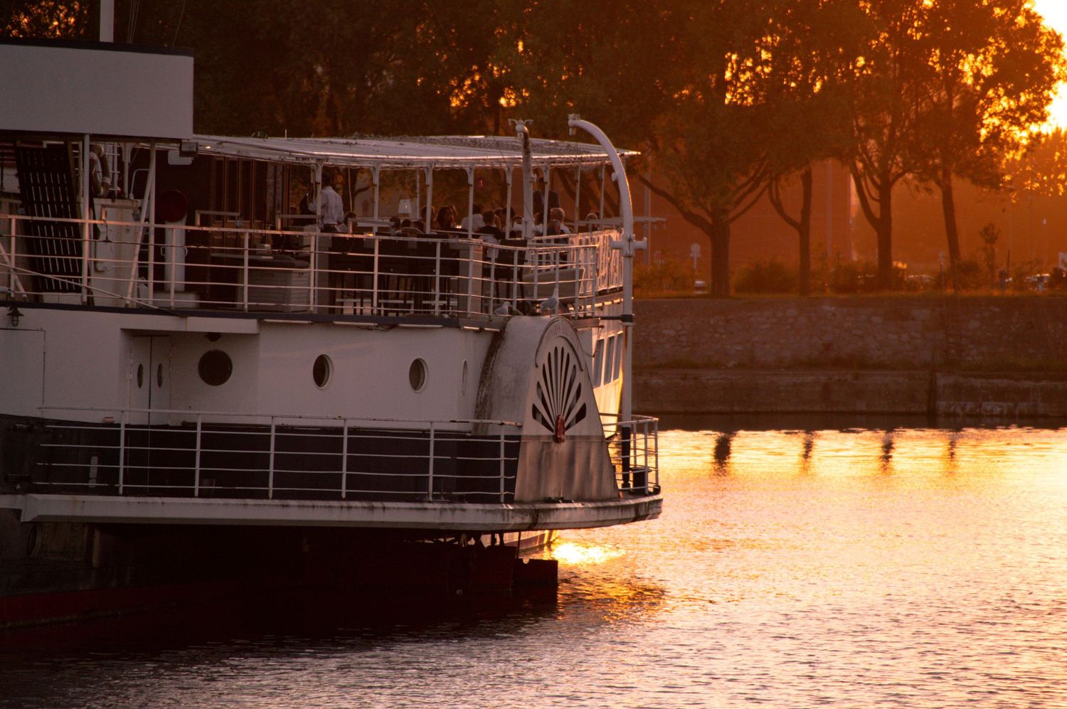 A riverboat cruise ship docked at sunset, with golden light reflecting on the water and silhouetted trees in the background.