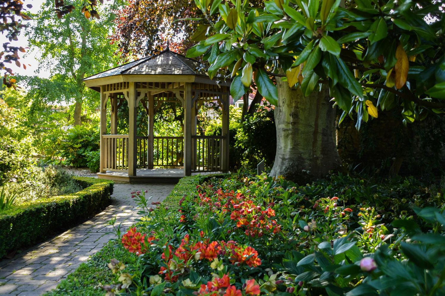 A scenic garden path lined with lush greenery and vibrant red flowers leads to a gazebo.