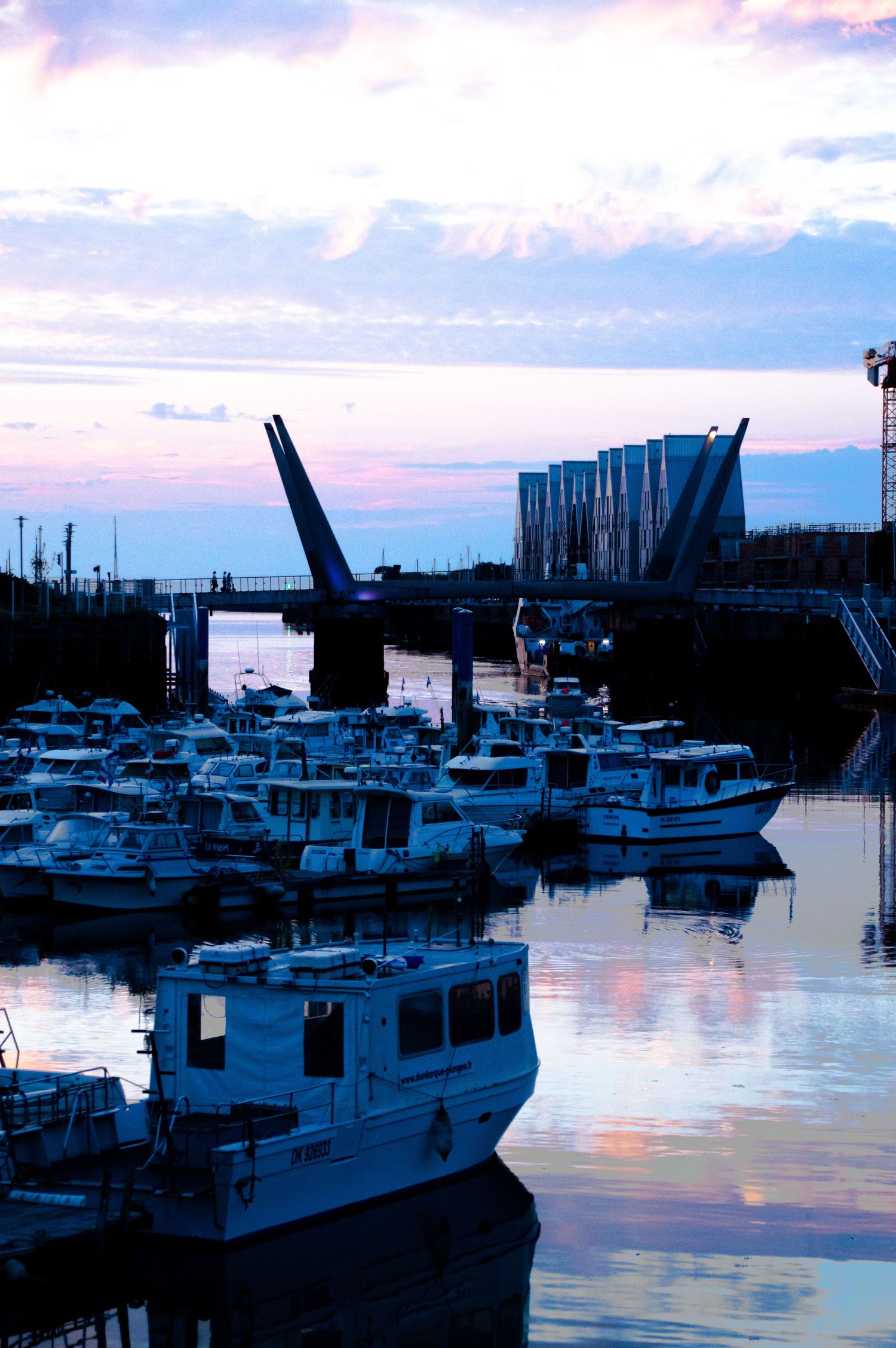 Boats docked in a marina at dusk with a drawbridge and colorful pink and blue sunset sky reflecting on calm water.