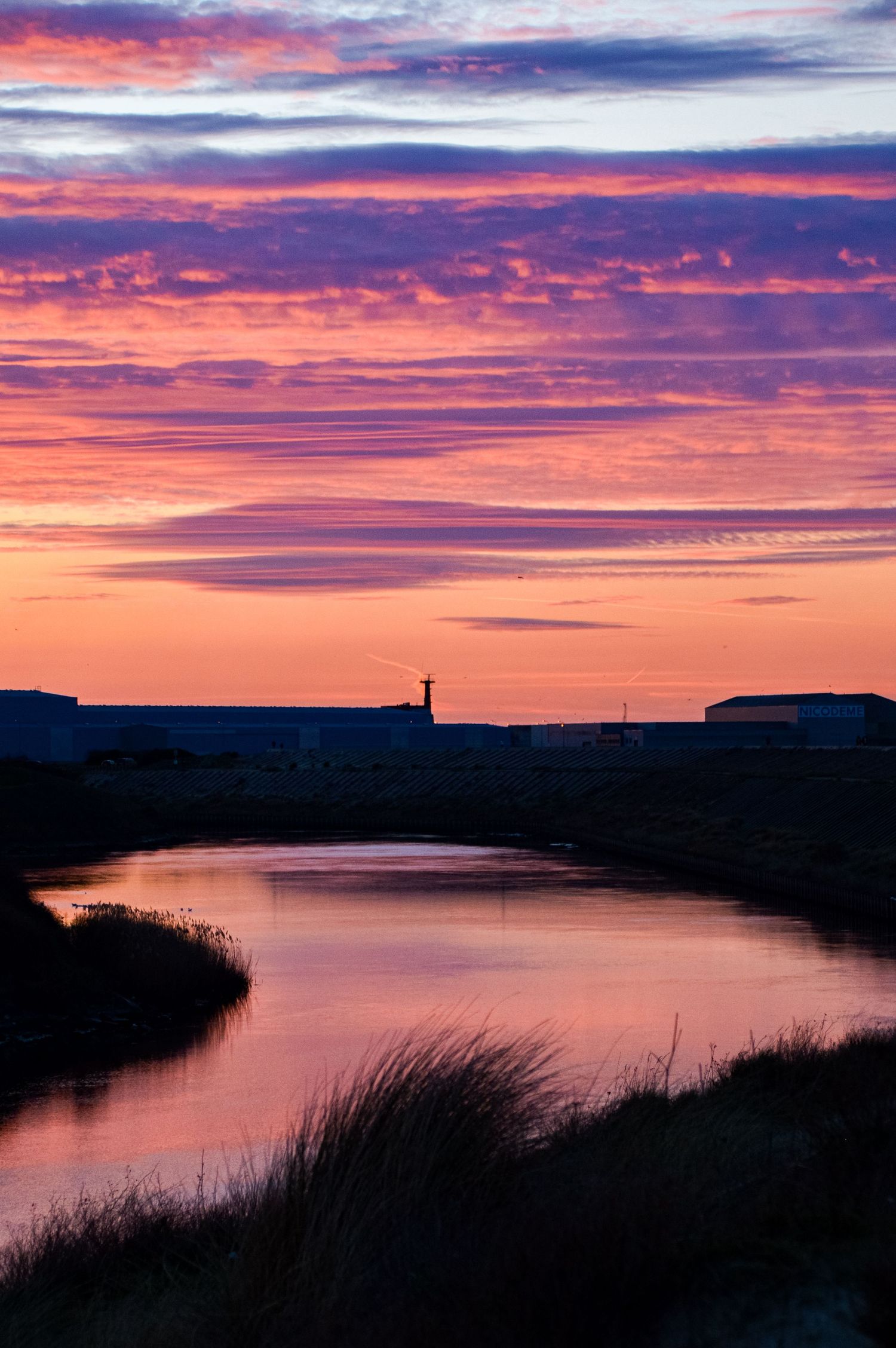 Harbor entrance with lighthouse at sunset reflecting pink and purple clouds over water.