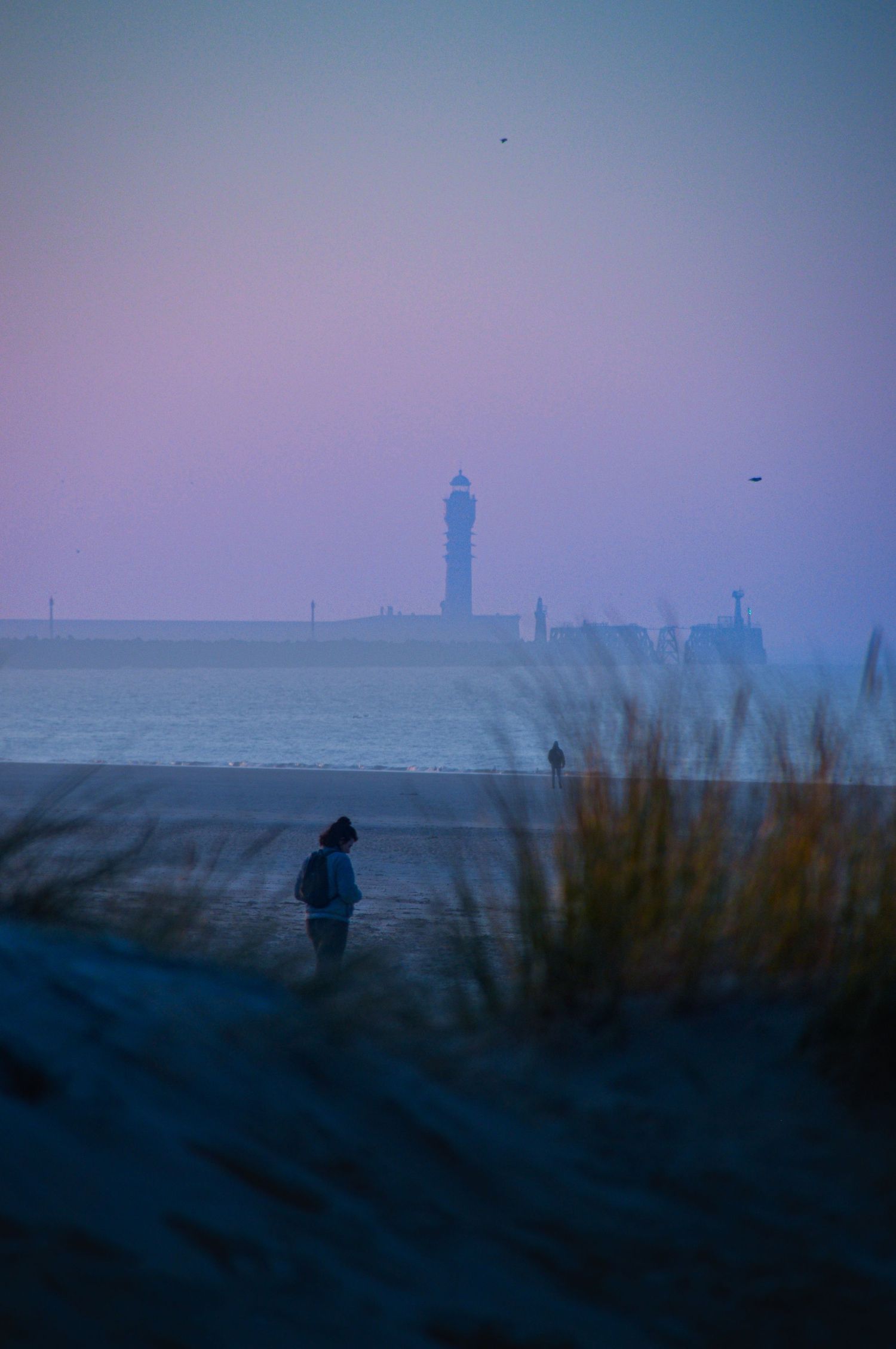 Silhouette solitaire sur la plage avec un phare (le feu de saint-pol à Dunkerque) se détachant sur un ciel violet de crépuscule.