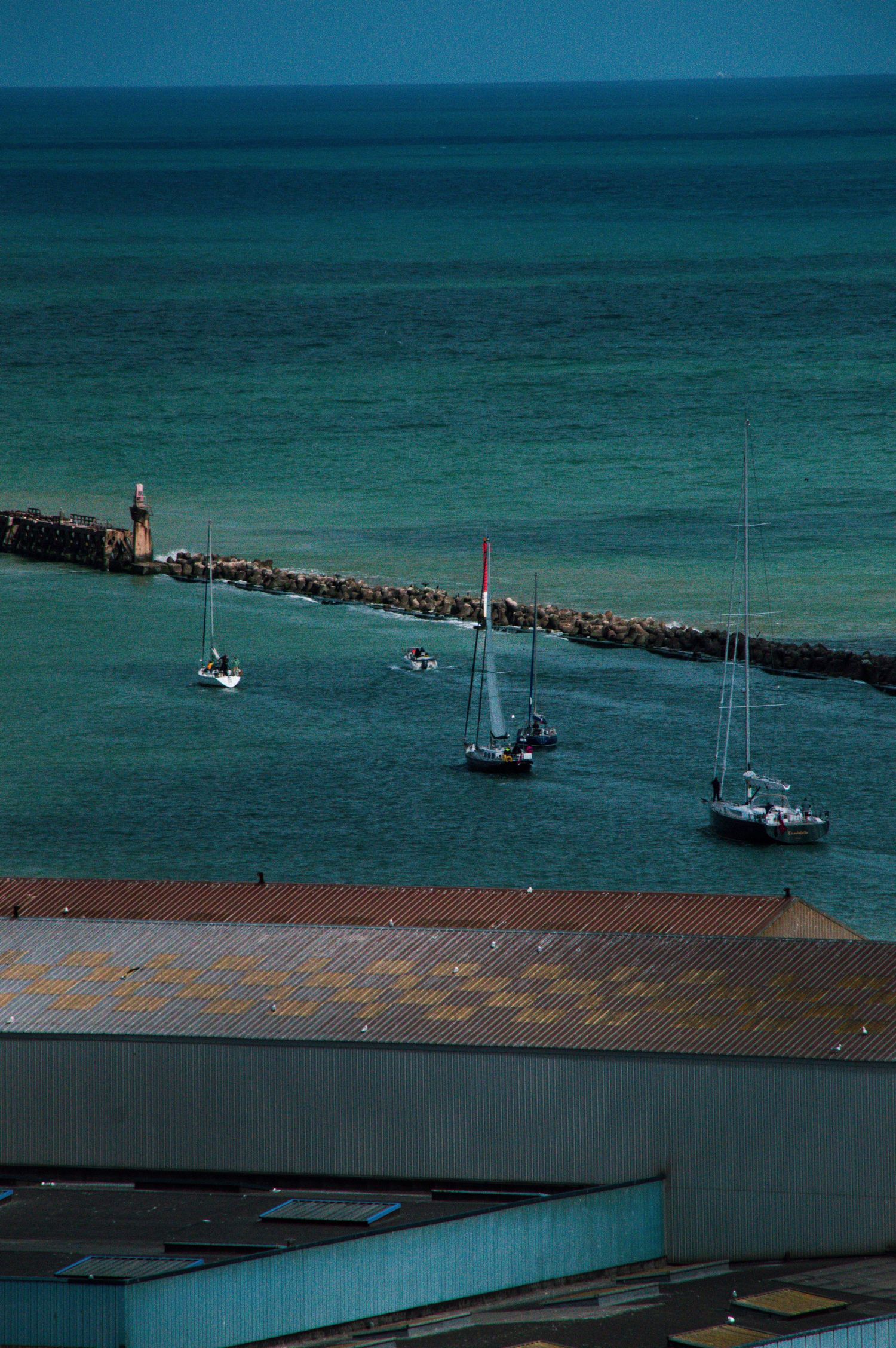 Aerial view of sailboats near a rocky breakwater in turquoise ocean waters, with a waterfront building roof in foreground.
